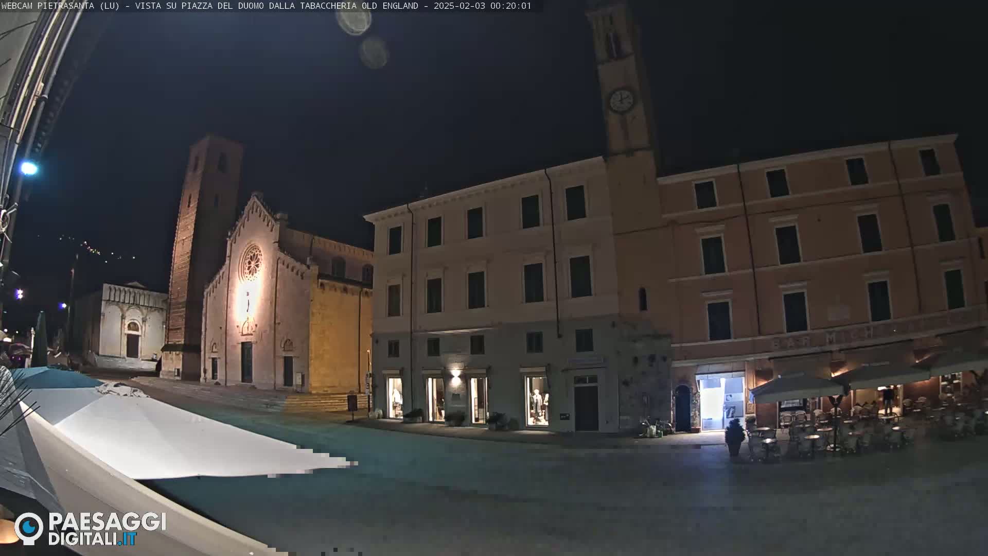 A nighttime view of a town square with illuminated buildings, including a church and a bar with outdoor seating, under a clear, dark sky.