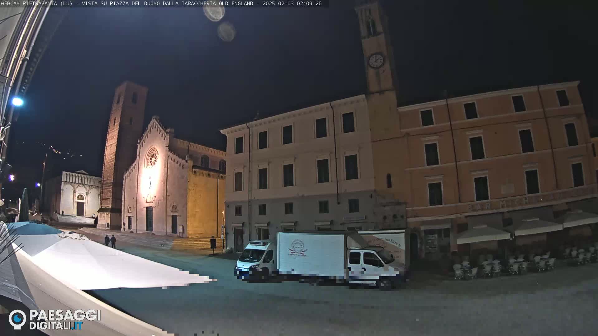 A nighttime view of a town square with a church, buildings, and a parked delivery truck under a clear, dark sky.