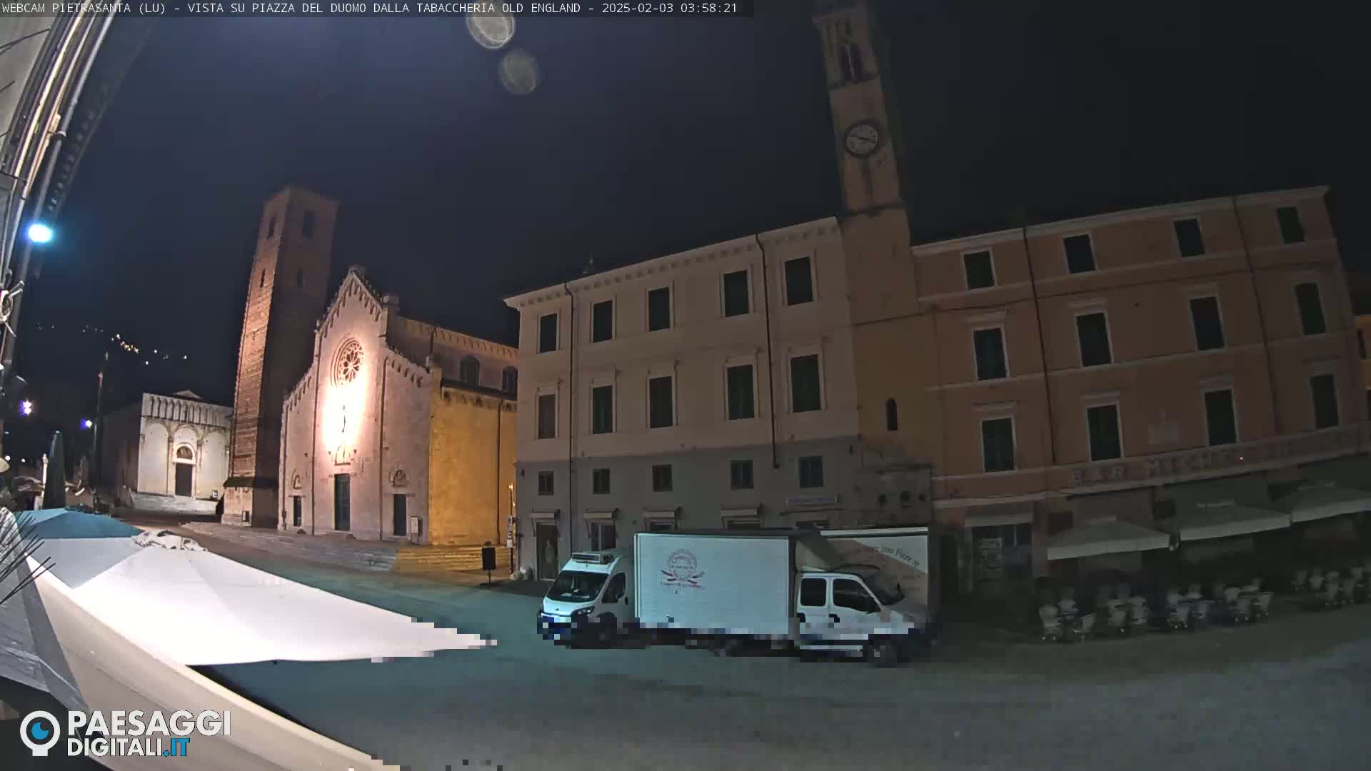A nighttime view of a town square, featuring a church, several buildings, a delivery truck, and outdoor seating areas under awnings.