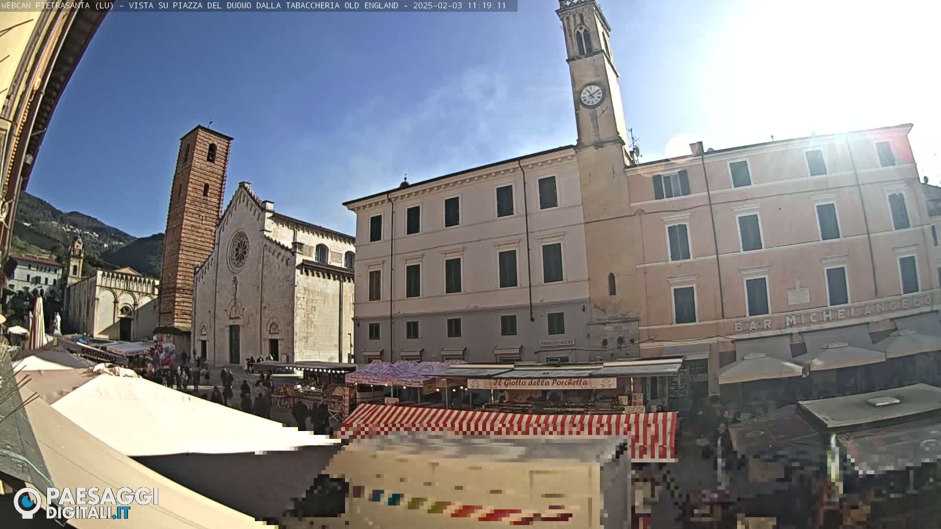 A sunny day in a European town square, showing a large church, market stalls, and several buildings, including a clock tower.
