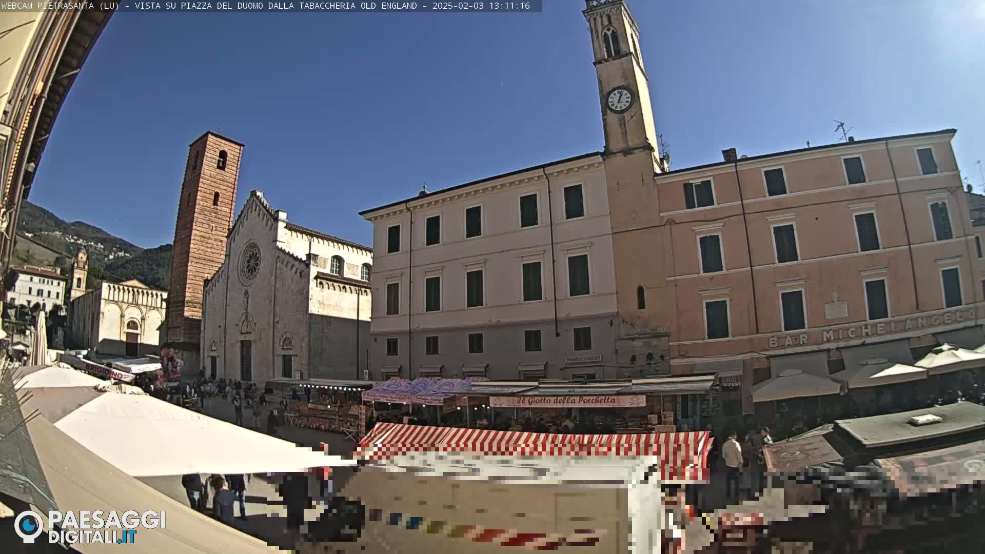 A sunny town square is bustling with an outdoor market set against the backdrop of historic buildings, including a tall church tower and a large church.