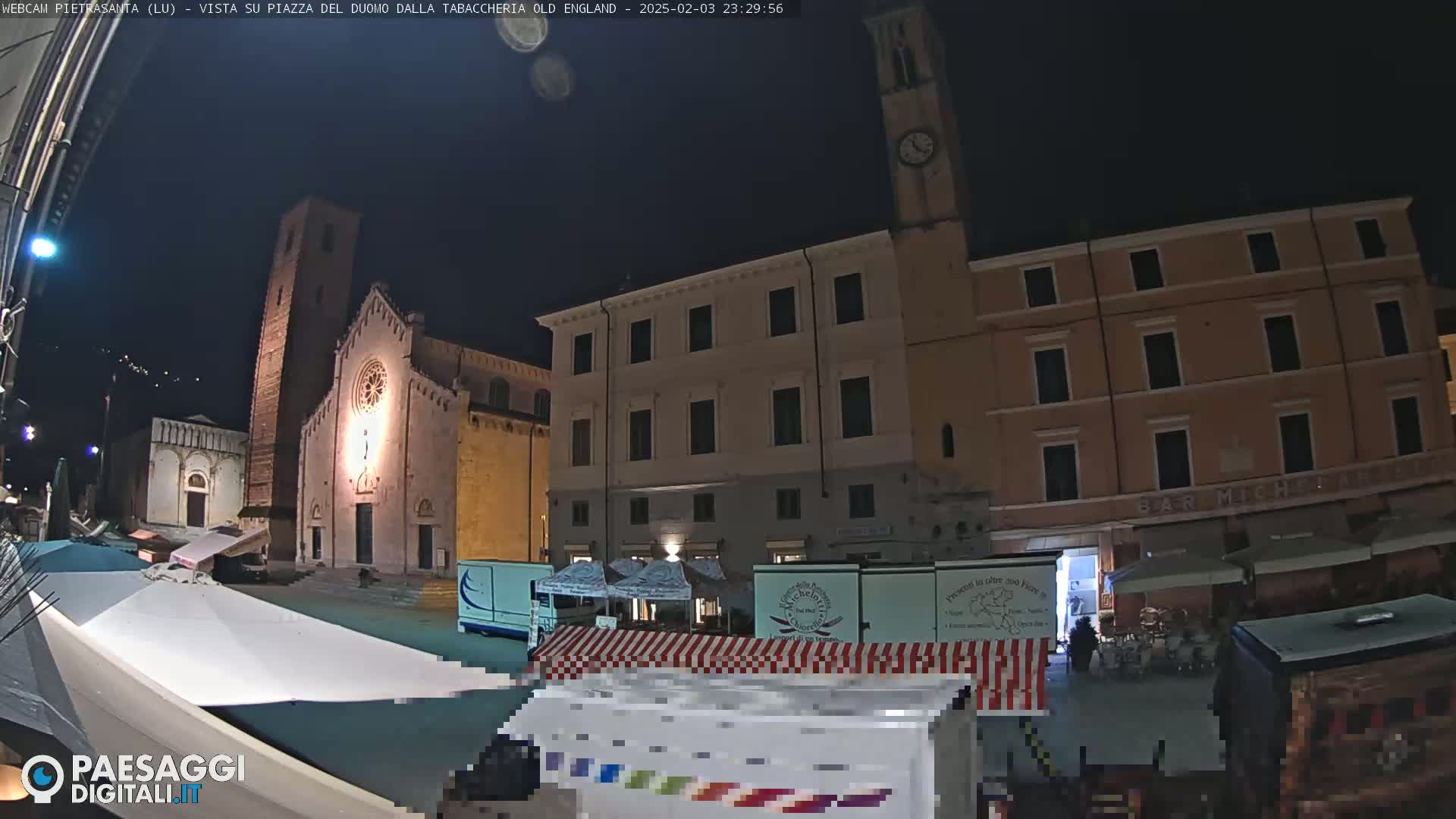 A nighttime view of a town square with several buildings, including a church and a tall clock tower, market stalls, and some people, under a clear, dark sky.