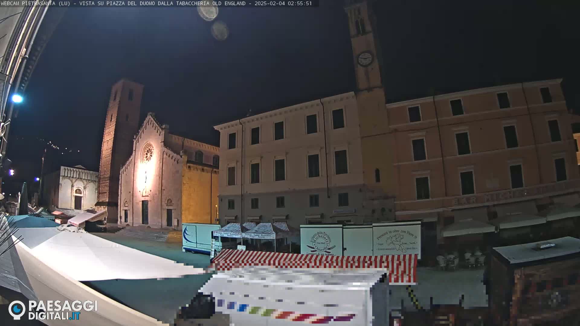 At night, a town square is sparsely populated with market stalls under a dark sky, illuminated by lights on buildings including a church and clock tower.
