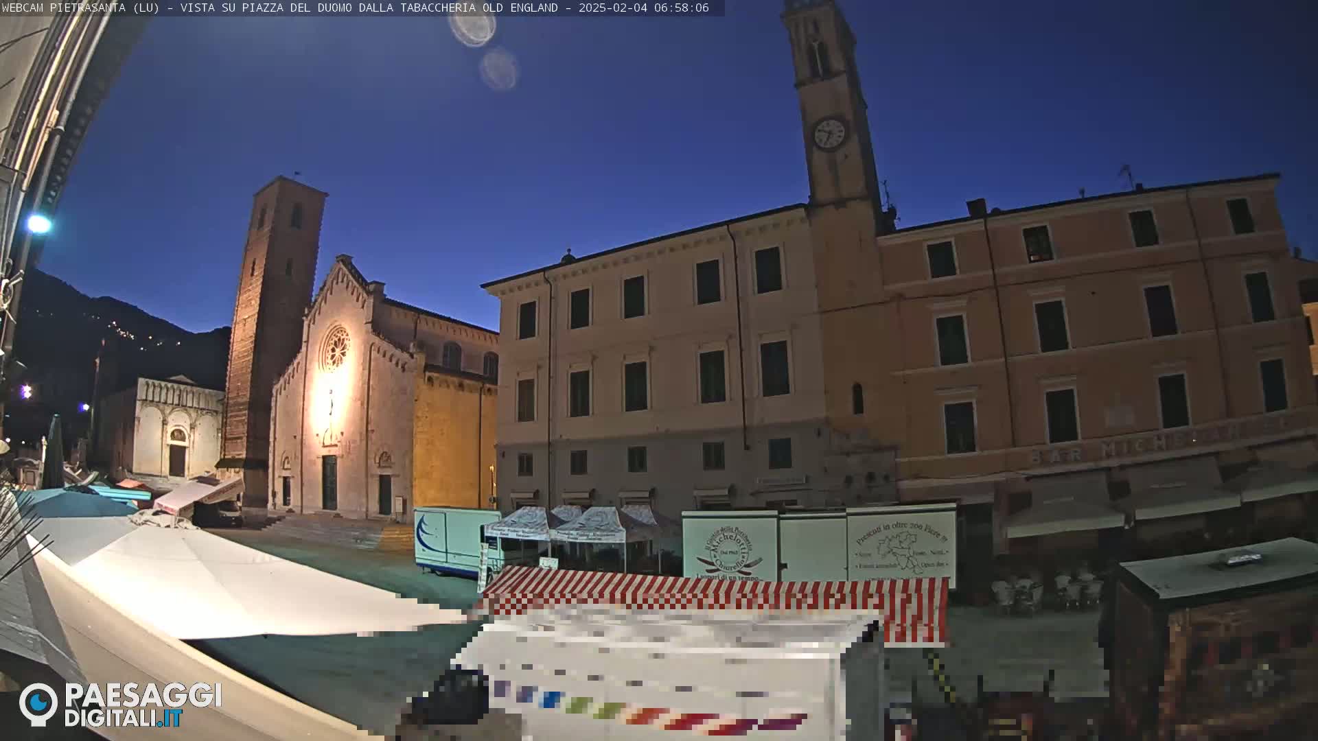 A twilight view of a town square with a church, buildings, and market stalls under a clear, dark blue sky.