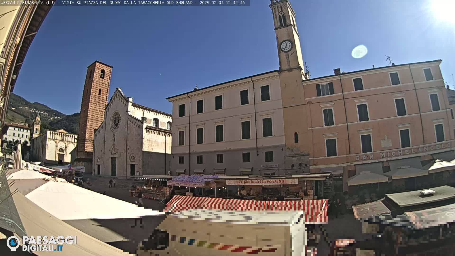 A sunny day in a town square with a large church, a clock tower, and several market stalls set up in front of buildings.
