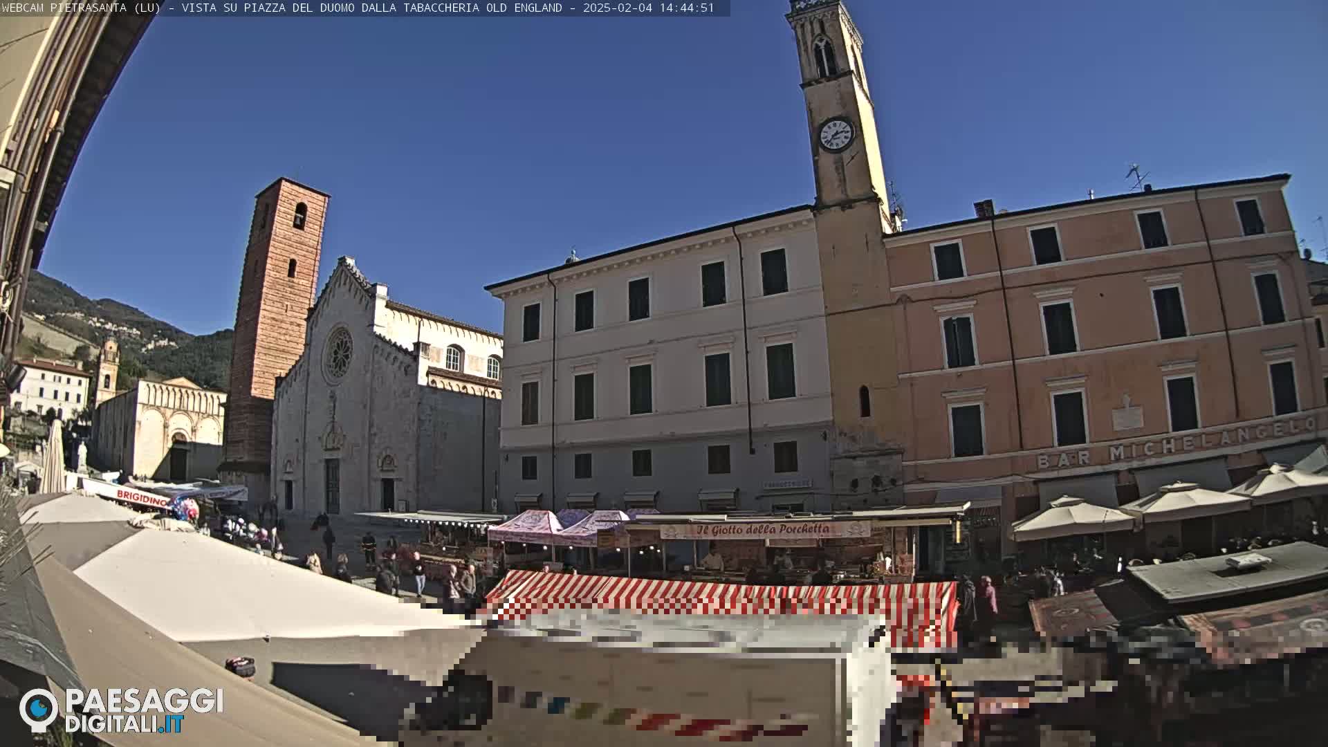 A sunny day in a town square with a large church, several buildings, and market stalls, partially obscured by awnings.
