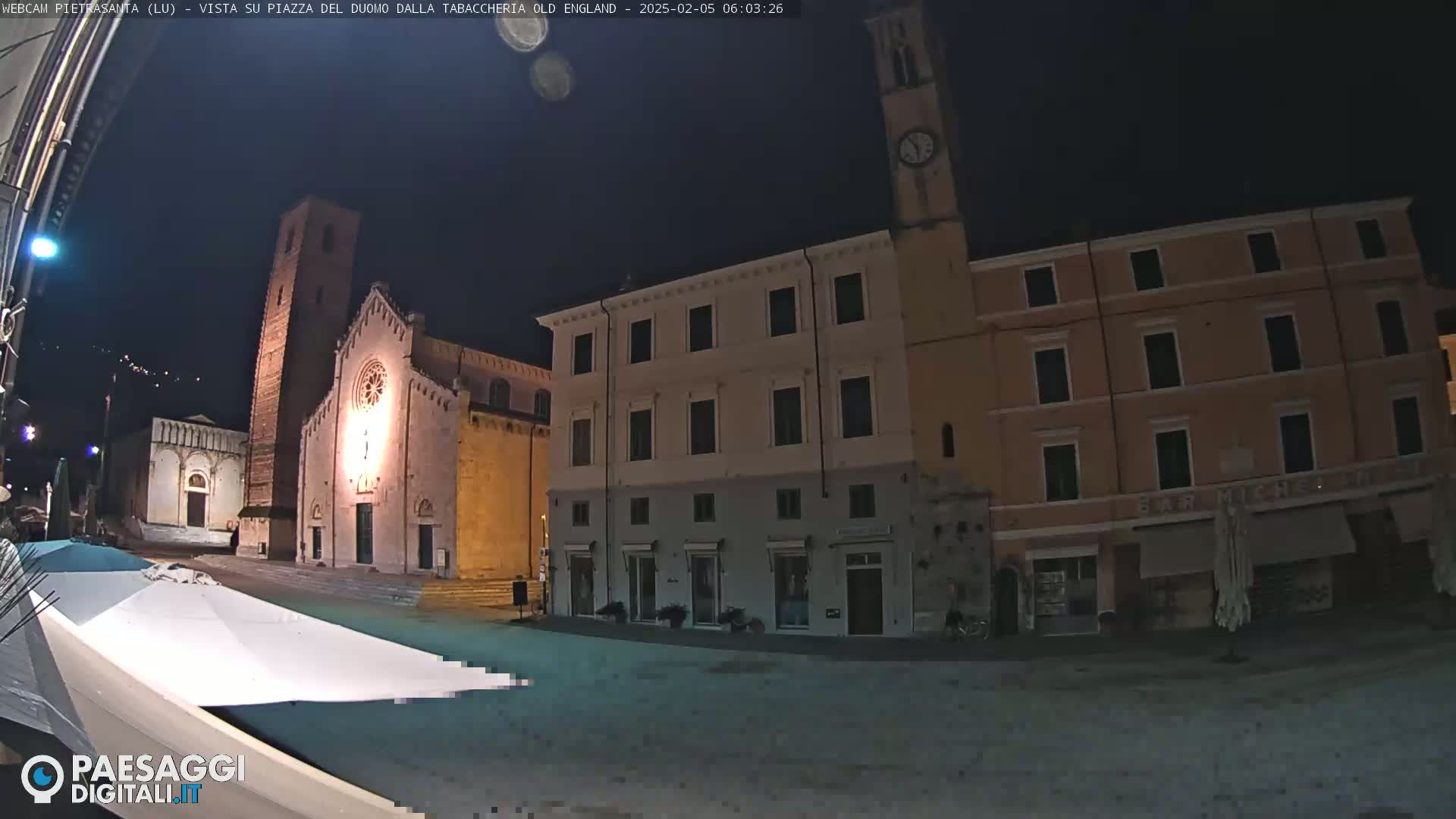 A nighttime view of a town square with several buildings, including a church with a tall bell tower, under a clear, dark sky.