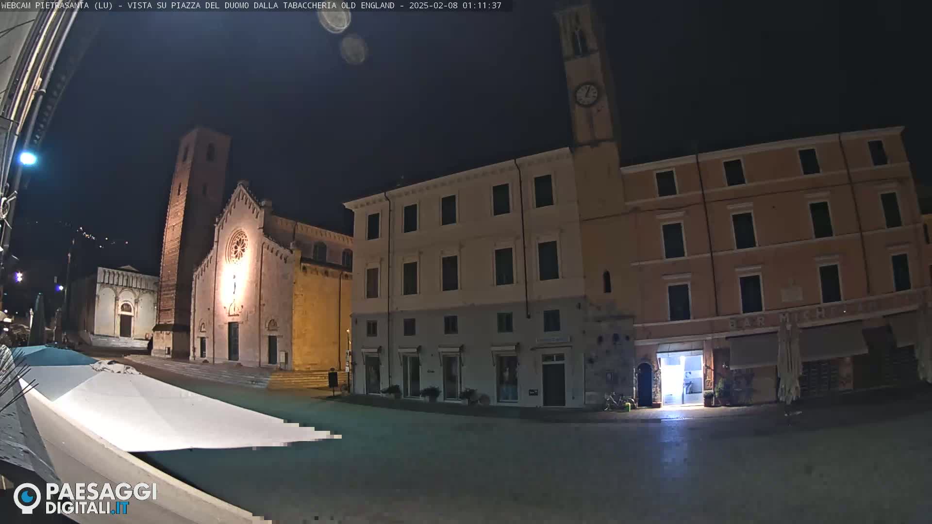 A nighttime, low-light view of a town square featuring a church with a tall bell tower, adjacent buildings, and a partially covered outdoor seating area.