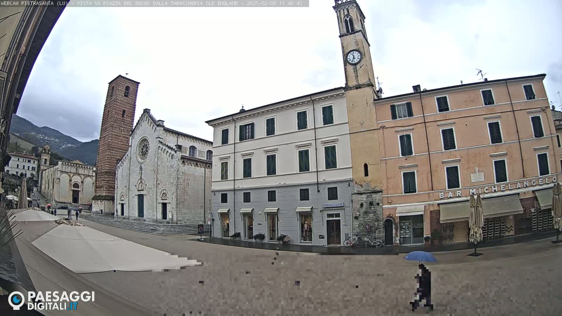 A town square with several buildings, including a church with a tall bell tower, under an overcast sky; a person with an umbrella is walking across the square.