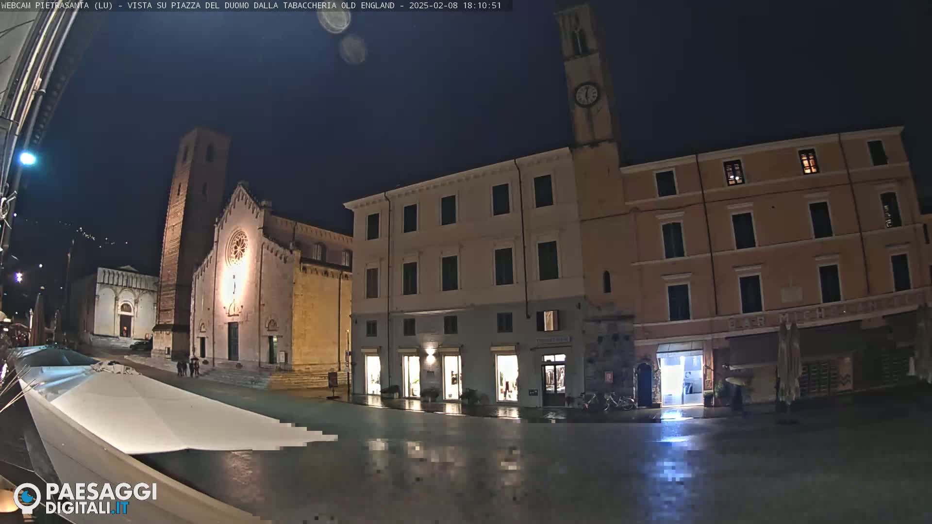 A wet, nighttime town square is shown with several buildings, including a church with a tall tower and clock tower, under a dark sky.
