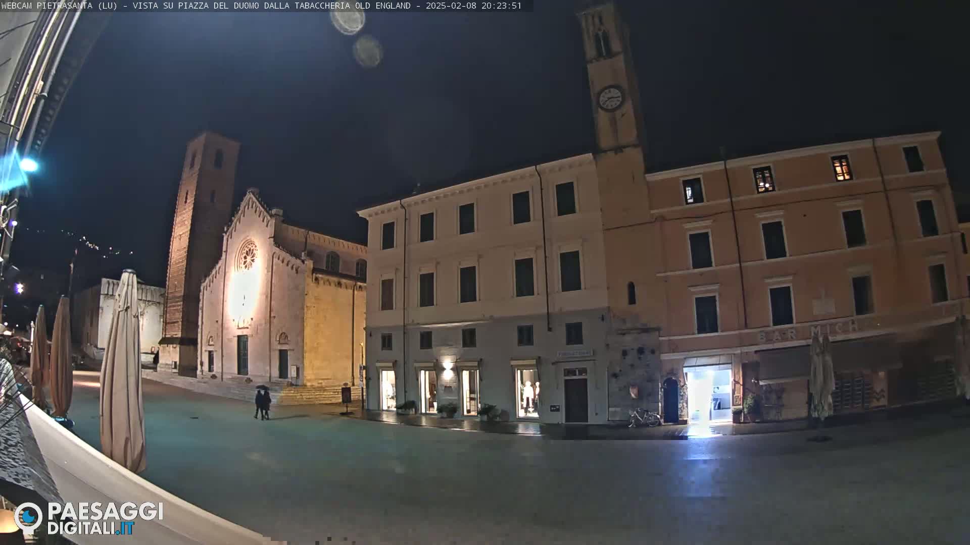 A nighttime view of a town square, featuring a church with a tall bell tower, several buildings, and a few people walking.