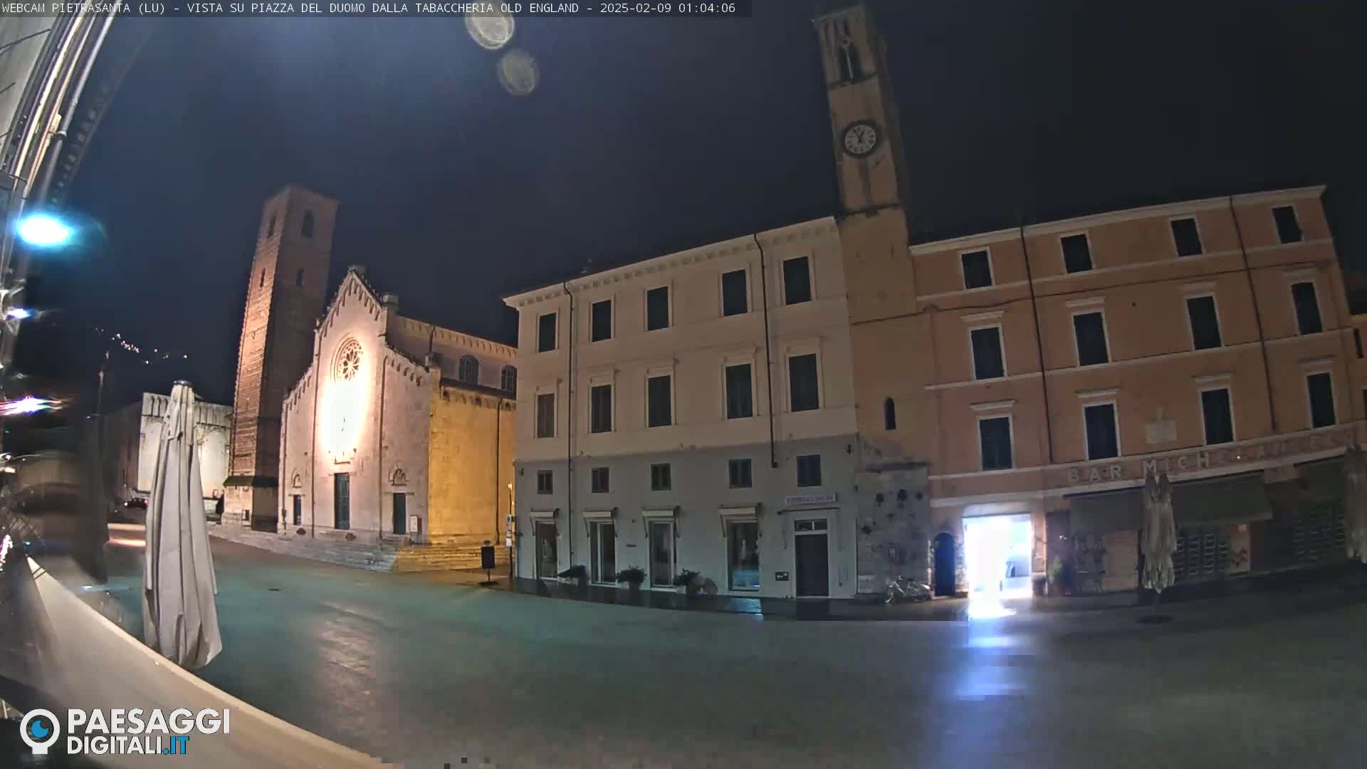 A nighttime view of a town square with illuminated buildings, including a church with a tall bell tower, under a dark sky.