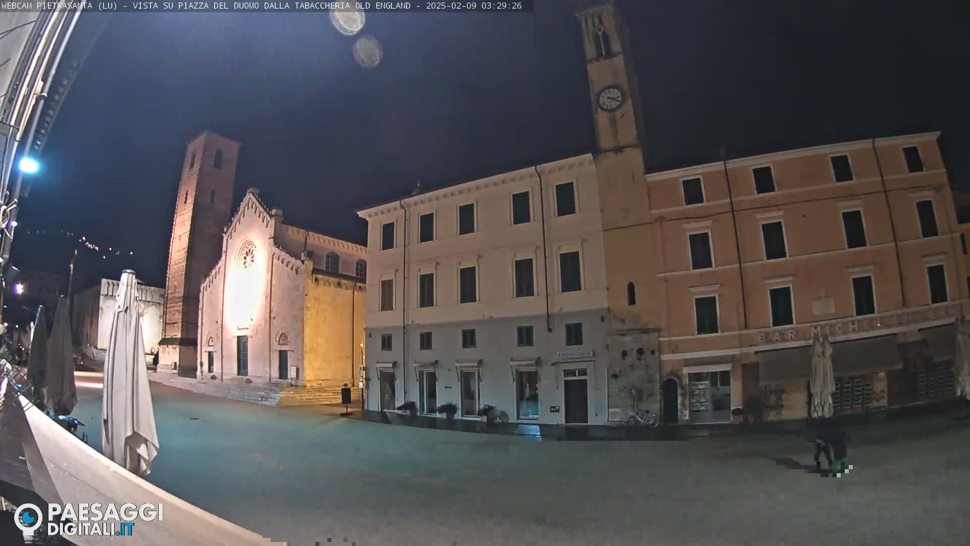 A nighttime view of a town square with several buildings, including a church with a tall tower, under a clear, dark sky.