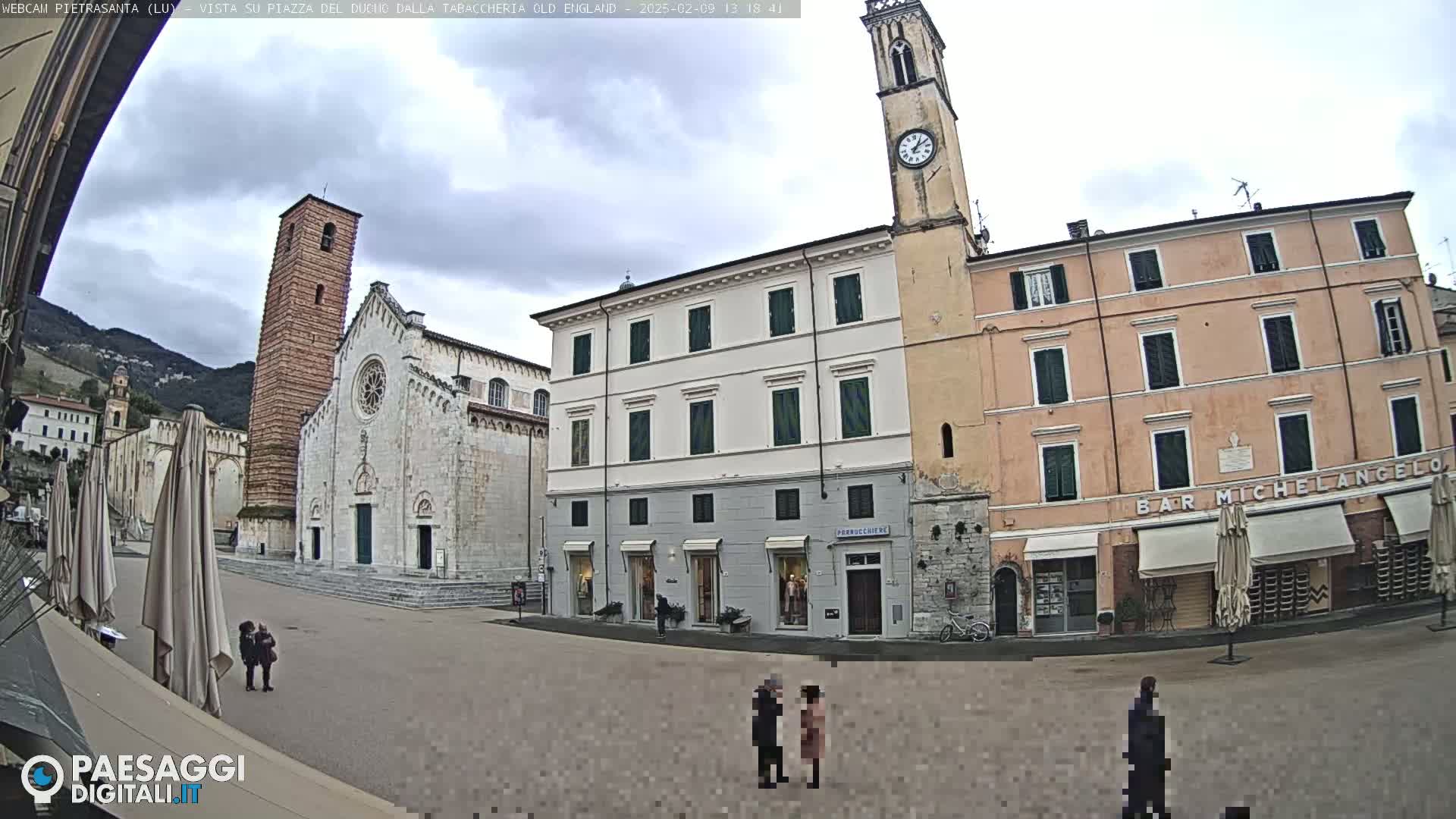 A town square with a church, clock tower, and several buildings under a cloudy sky.
