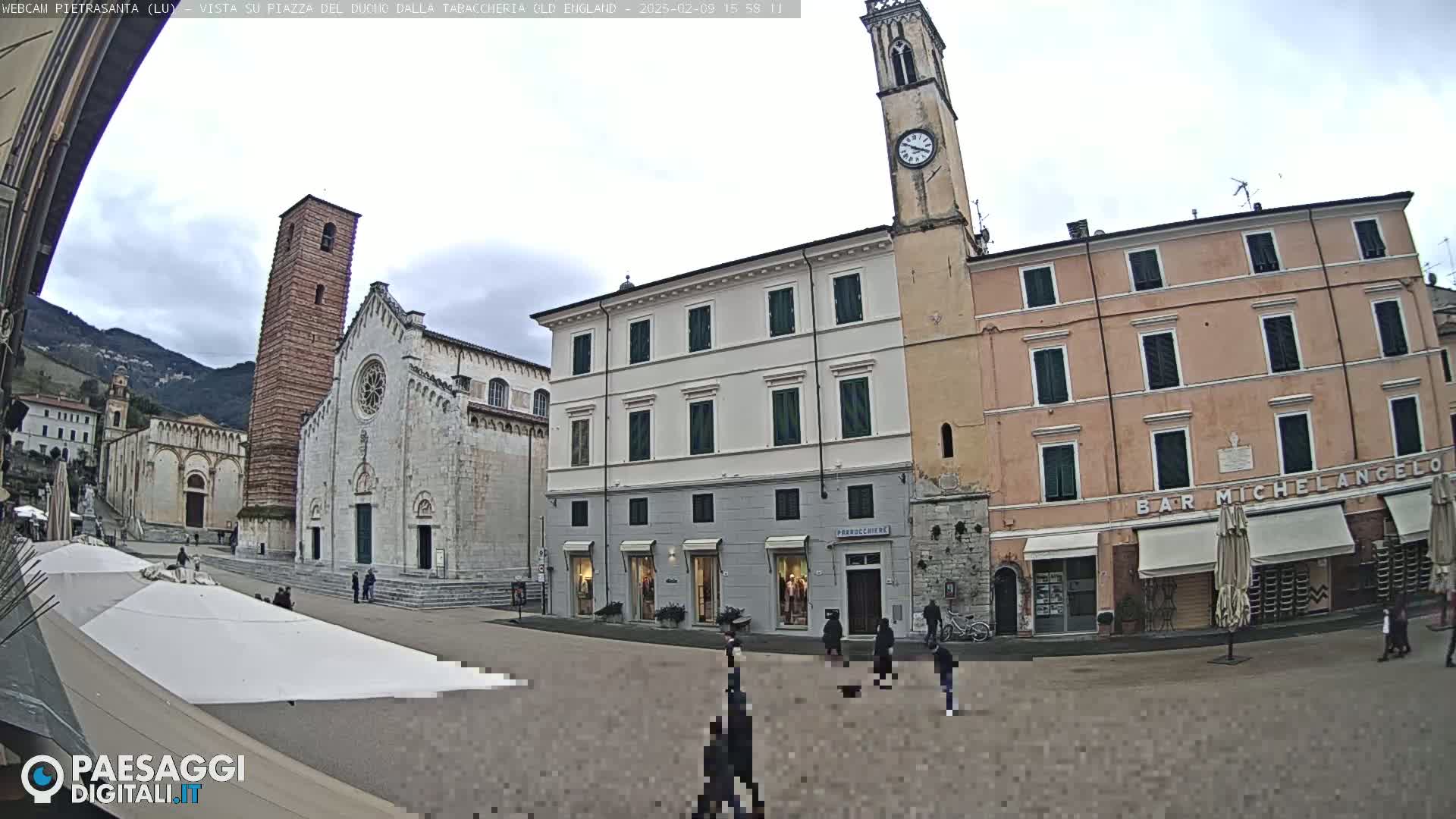 A town square is shown on an overcast day, with a church, clock tower, and several buildings surrounding it; several people are walking in the square.