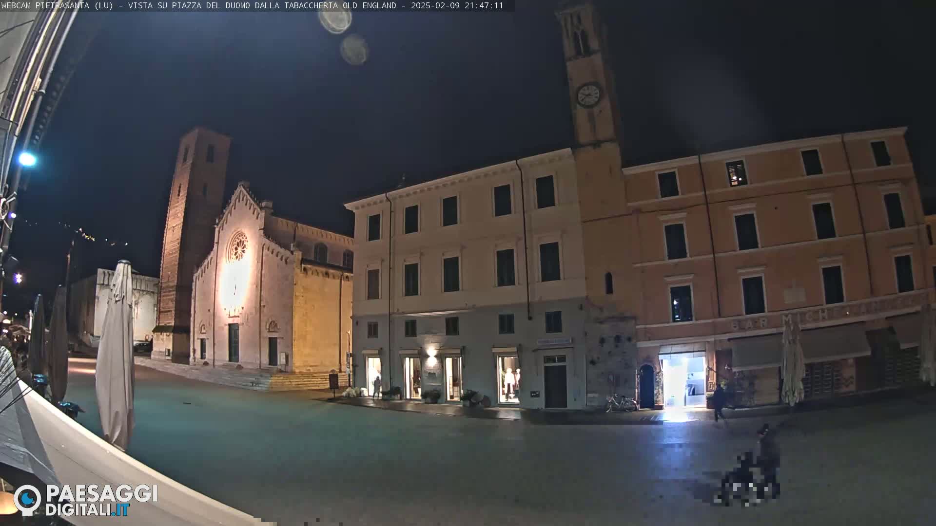 A nighttime view of a town square with a church, several buildings, and a few people, under a clear, dark sky.