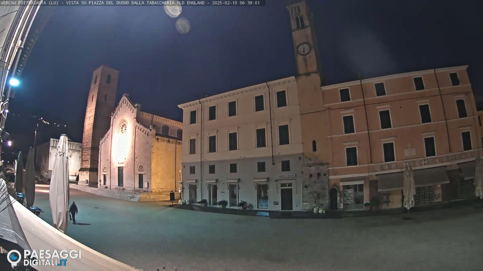 A nighttime, outdoor view of a town square with a church, buildings, and a person walking.