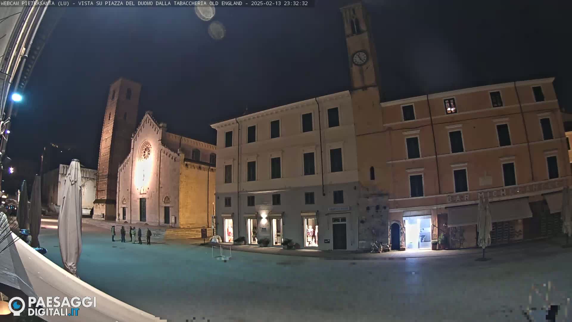 A nighttime view of a town square with a church, buildings, and a few people, under a clear, dark sky.