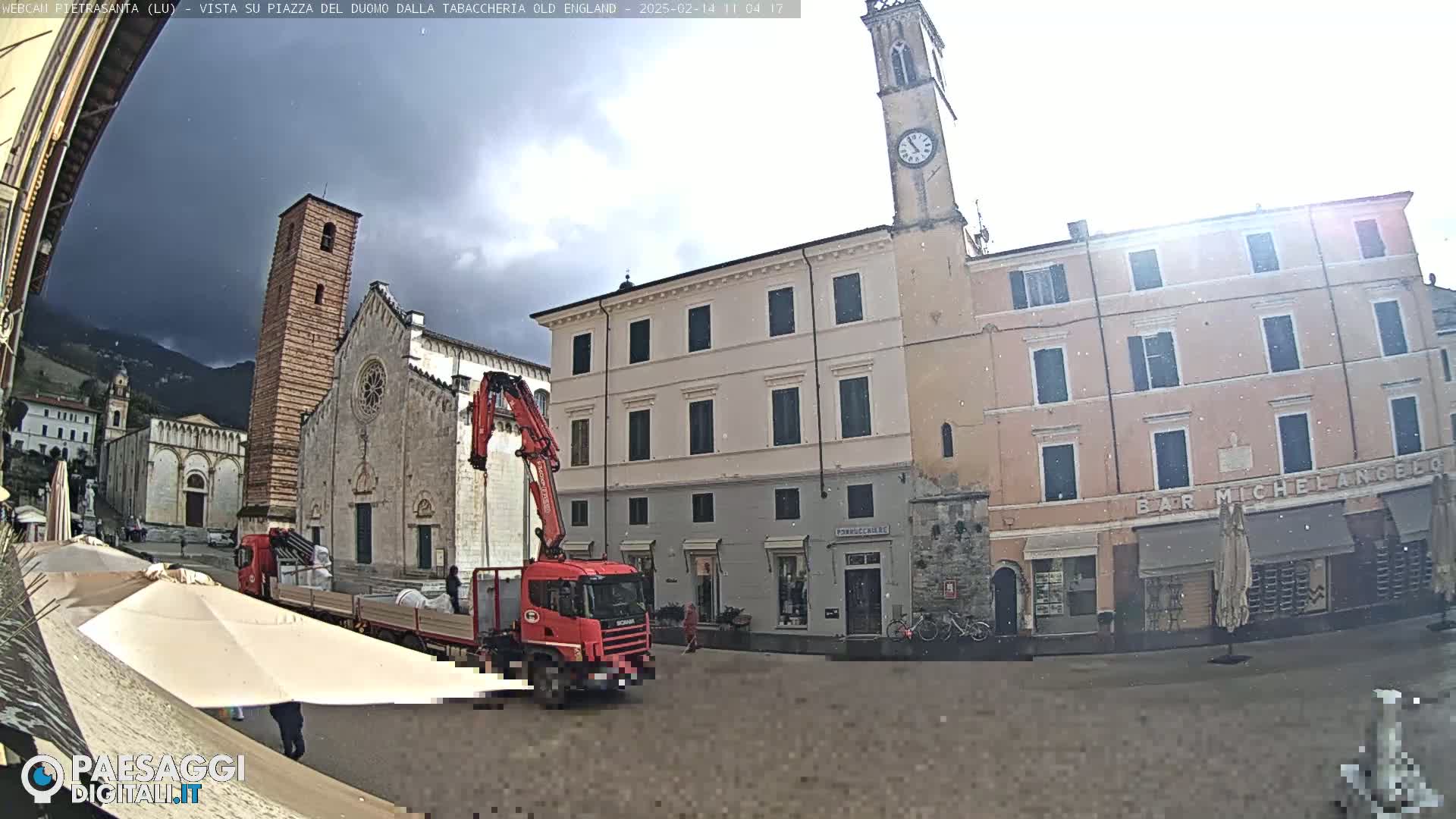 A town square with a church, clock tower, and buildings is partially obscured by a crane and truck unloading material on a somewhat overcast day with light precipitation.