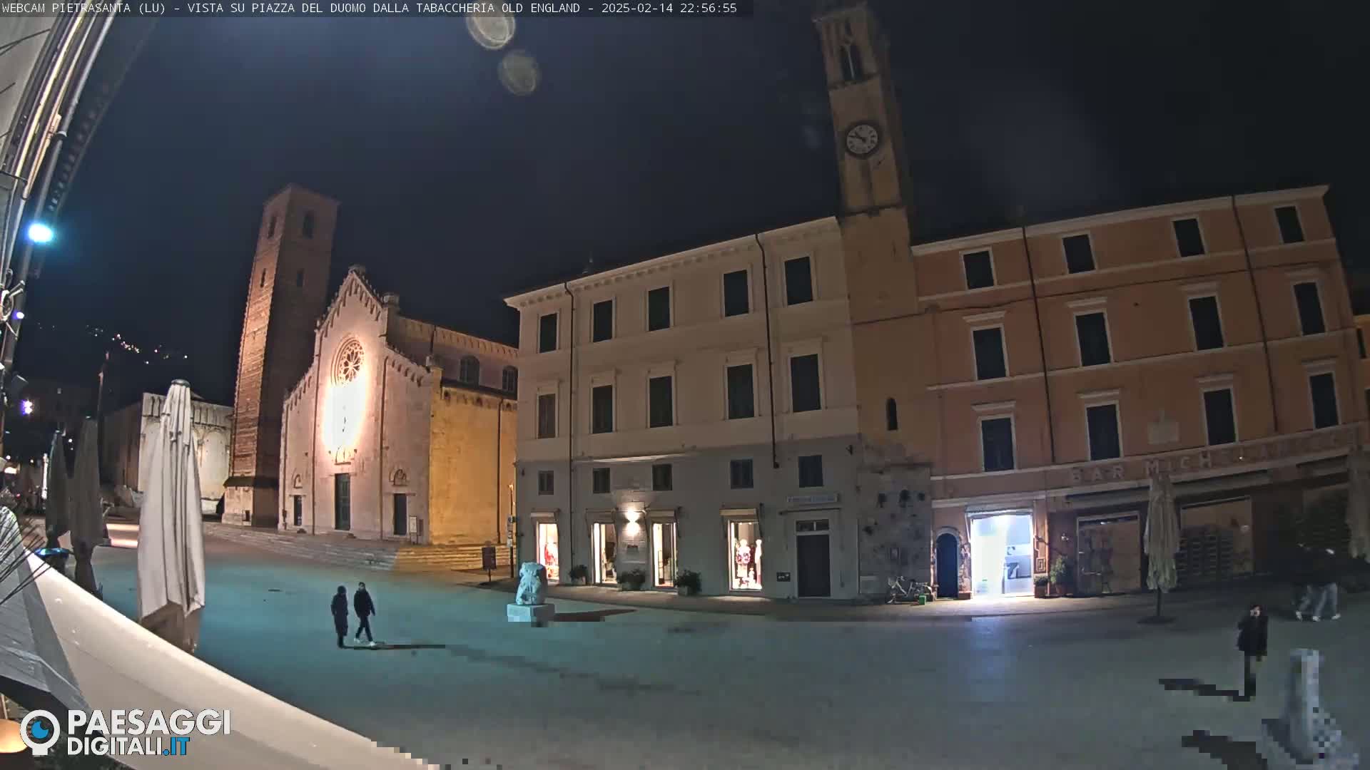 A nighttime view of a town square with a church, several buildings, and a few pedestrians under a clear, dark sky.