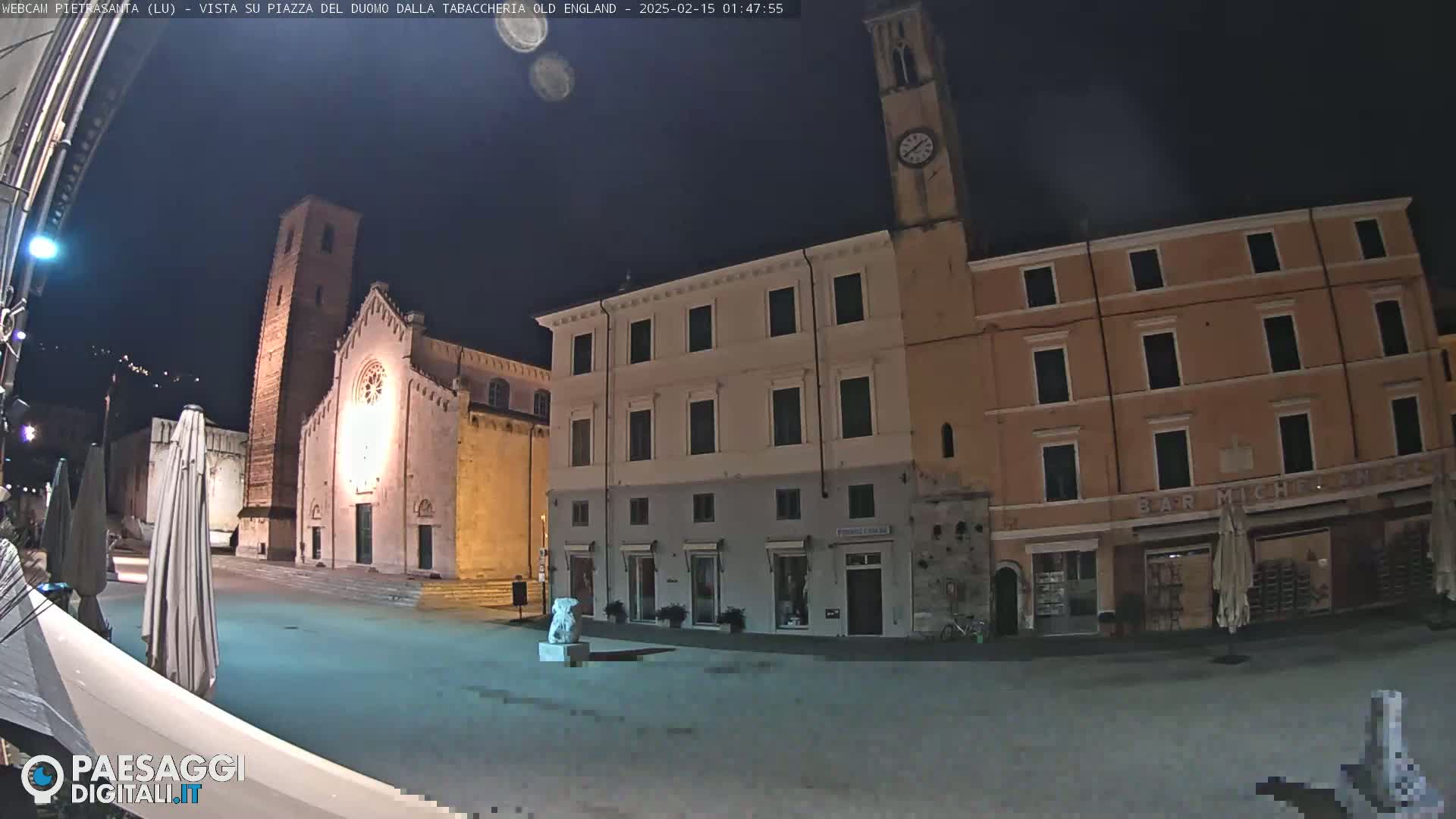 A nighttime view of a town square with illuminated buildings, including a church with a tall tower and a clock tower, under a dark sky.