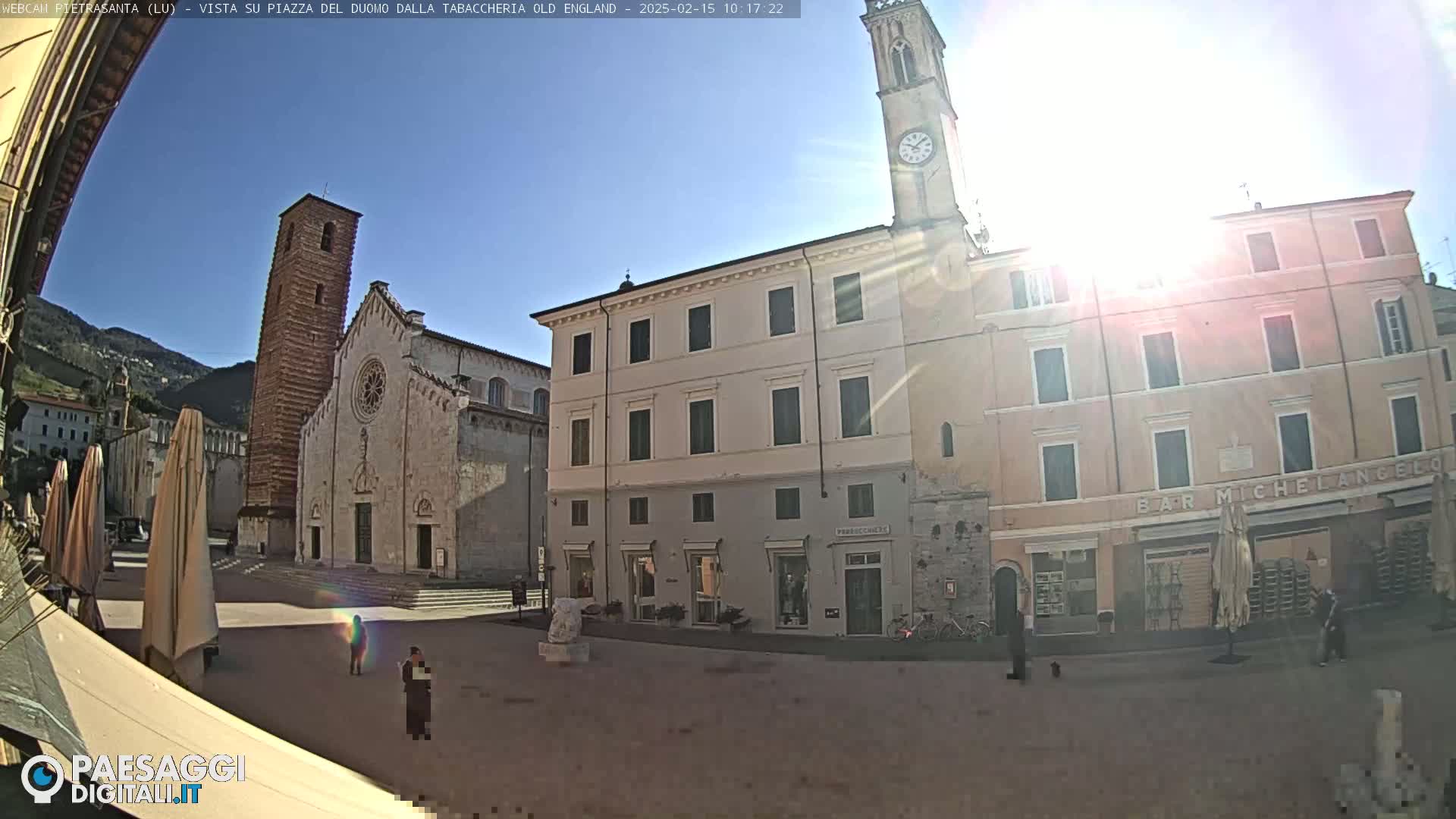 A sunny day in a town square, showing a large cathedral, several buildings, and a few people walking around.