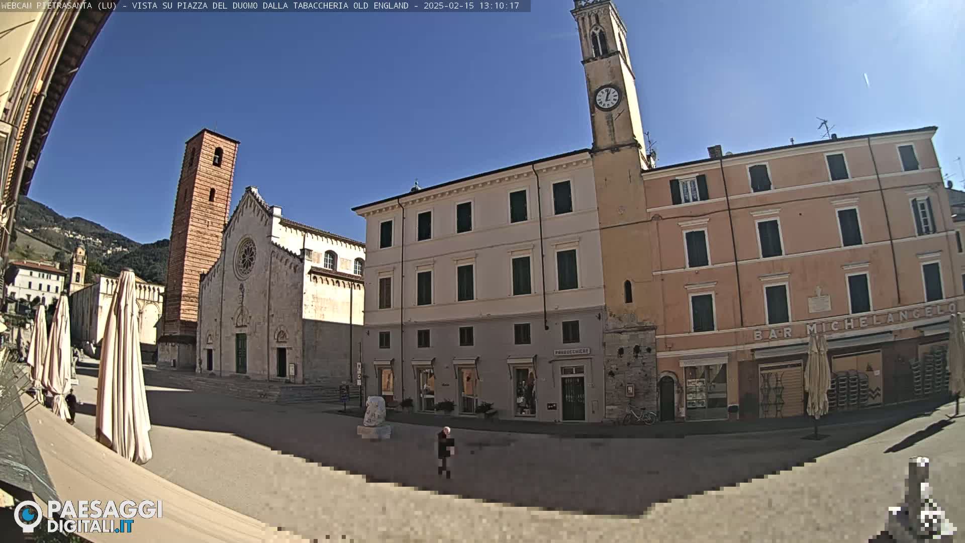 A sunny town square is seen, featuring a large church with a tall bell tower, several multi-story buildings, and a few people walking around.