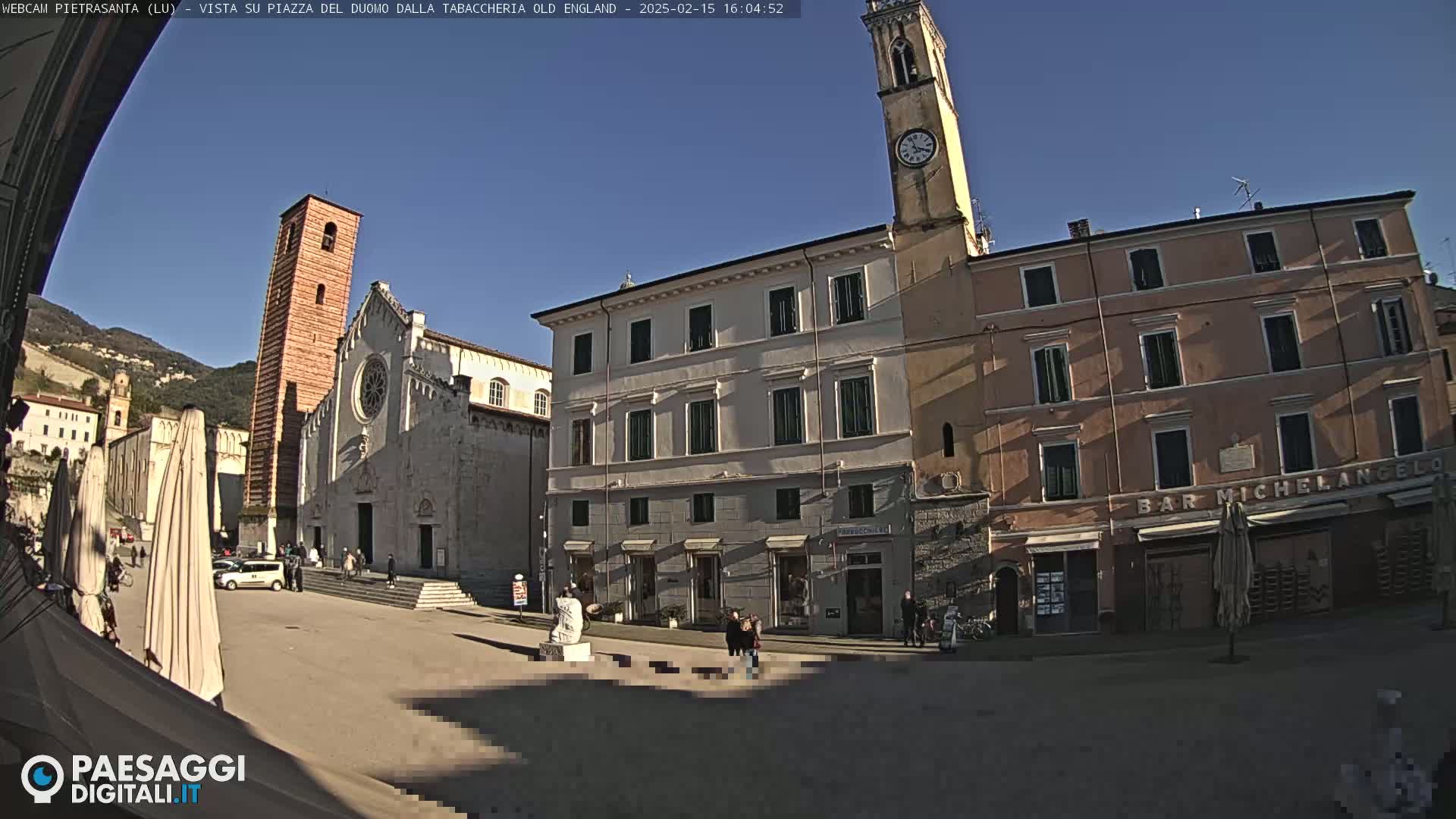 A town square, under sunny skies, features a large church with a tall bell tower, several buildings, and a few people.