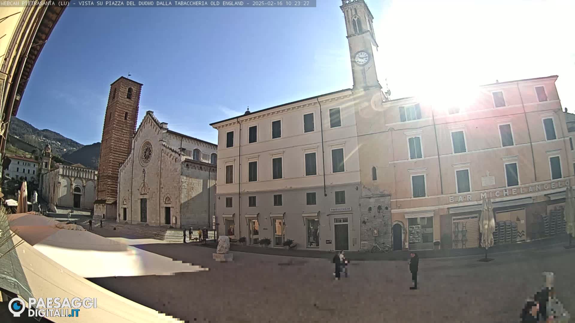 A sunny town square features a large cathedral, several multi-story buildings, and a few pedestrians.