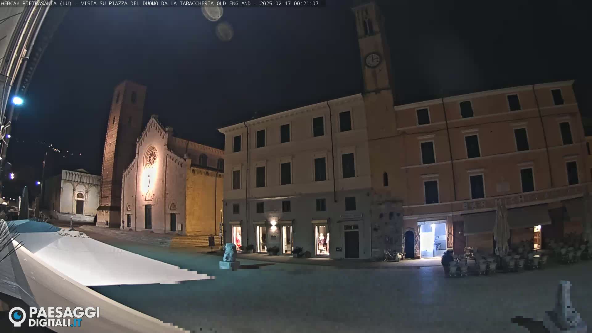 A nighttime view of a town square with a church, several buildings, and a partially covered outdoor seating area.