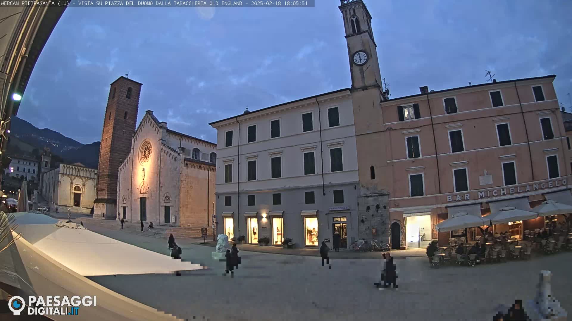 A town square at dusk, featuring a large church and several buildings, with a few people and outdoor seating visible under a twilight sky.