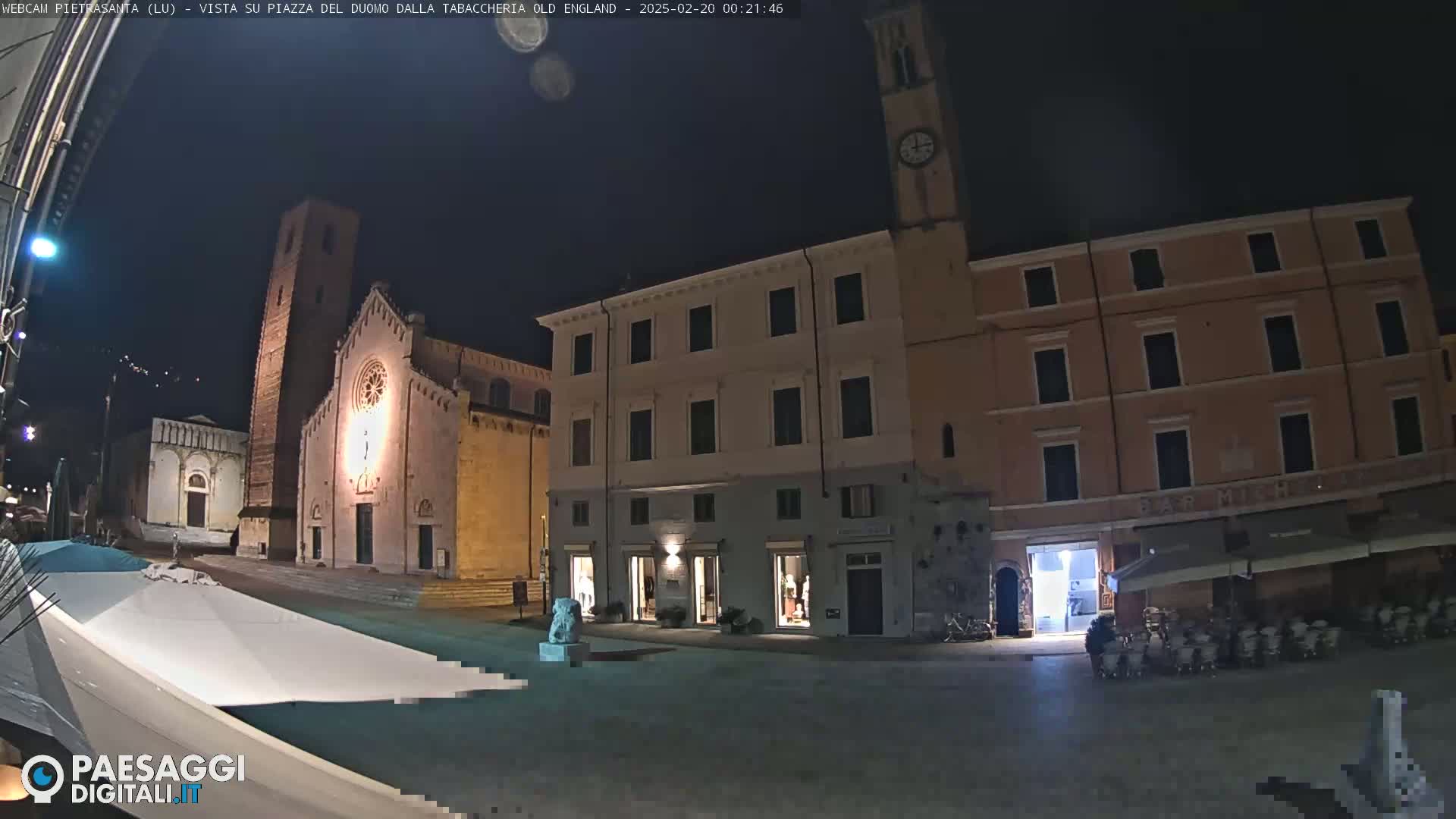 A nighttime view of a town square with a church, buildings, and outdoor seating areas under a clear sky.