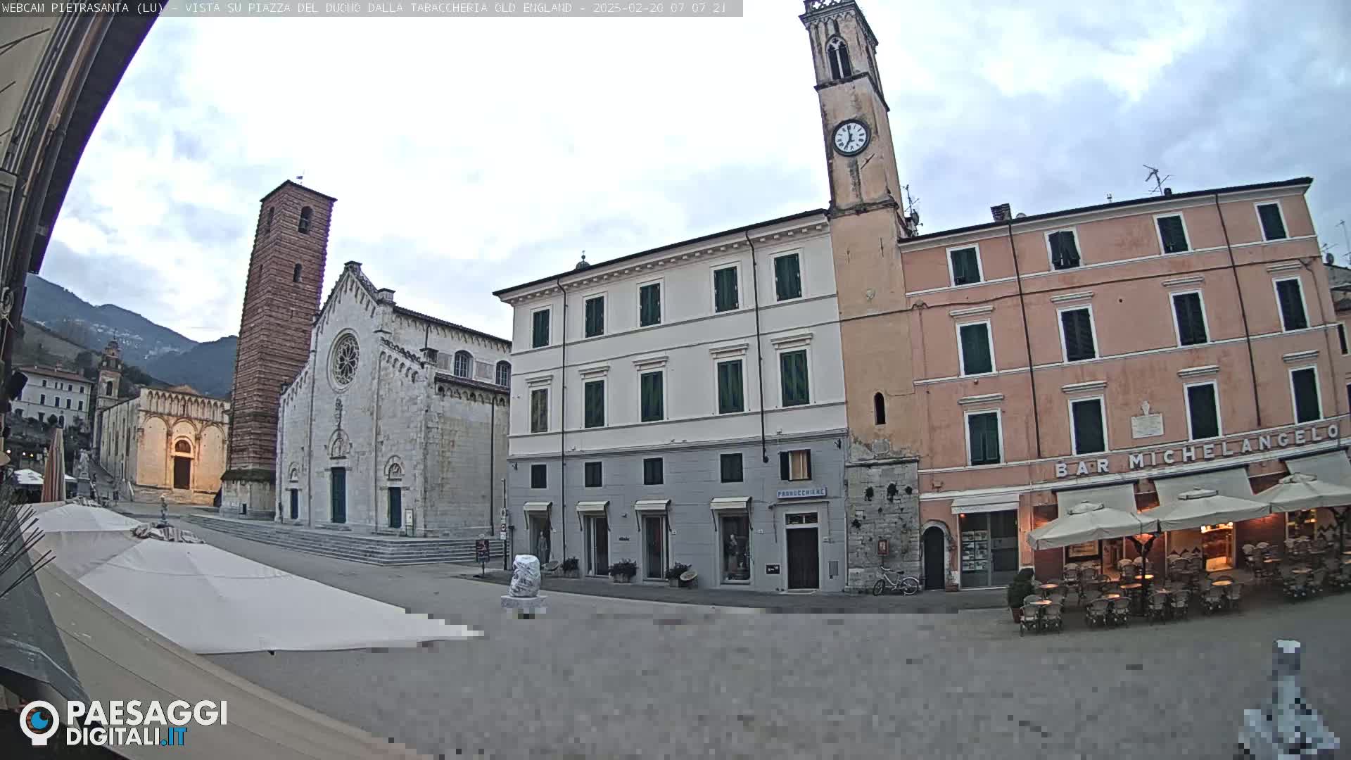A town square is shown on an overcast day, with a church, buildings, and outdoor seating visible.