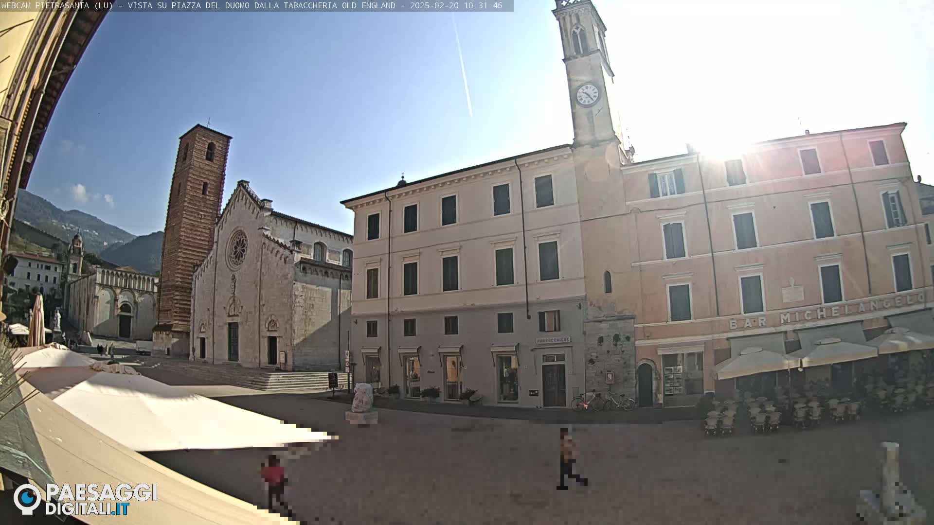 A sunny town square with a large cathedral, several buildings, and outdoor seating at a cafe, all situated against a backdrop of mountains.