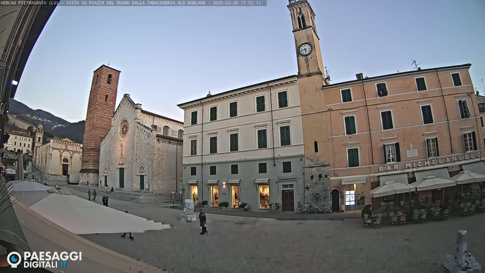 A town square in the late afternoon, under a clear sky, features a large church with a tall bell tower, several buildings, and outdoor seating at a cafe.