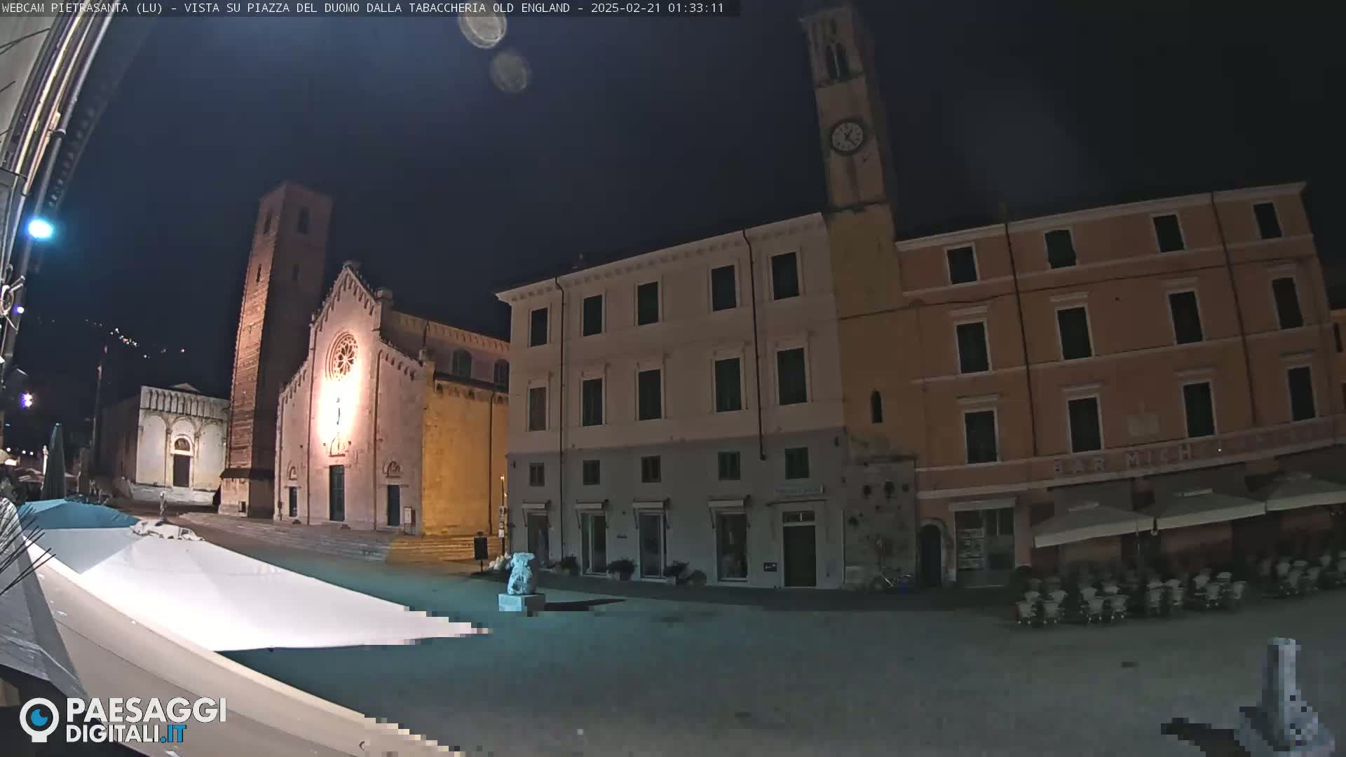 A nighttime view of a town square with a church, several buildings, and outdoor seating.