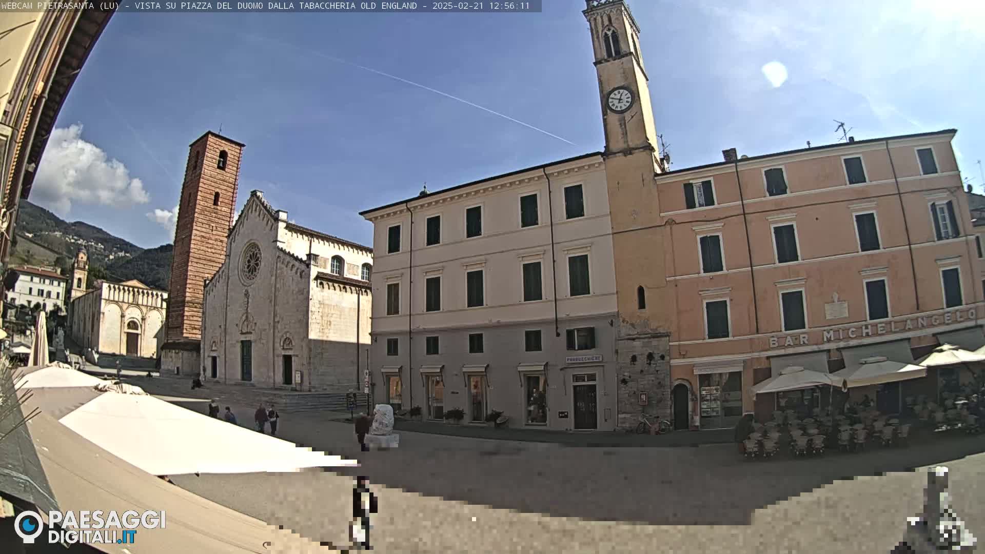 A sunny day reveals a town square in Italy, featuring a large cathedral, a clock tower, and several buildings, with outdoor seating at a cafe.