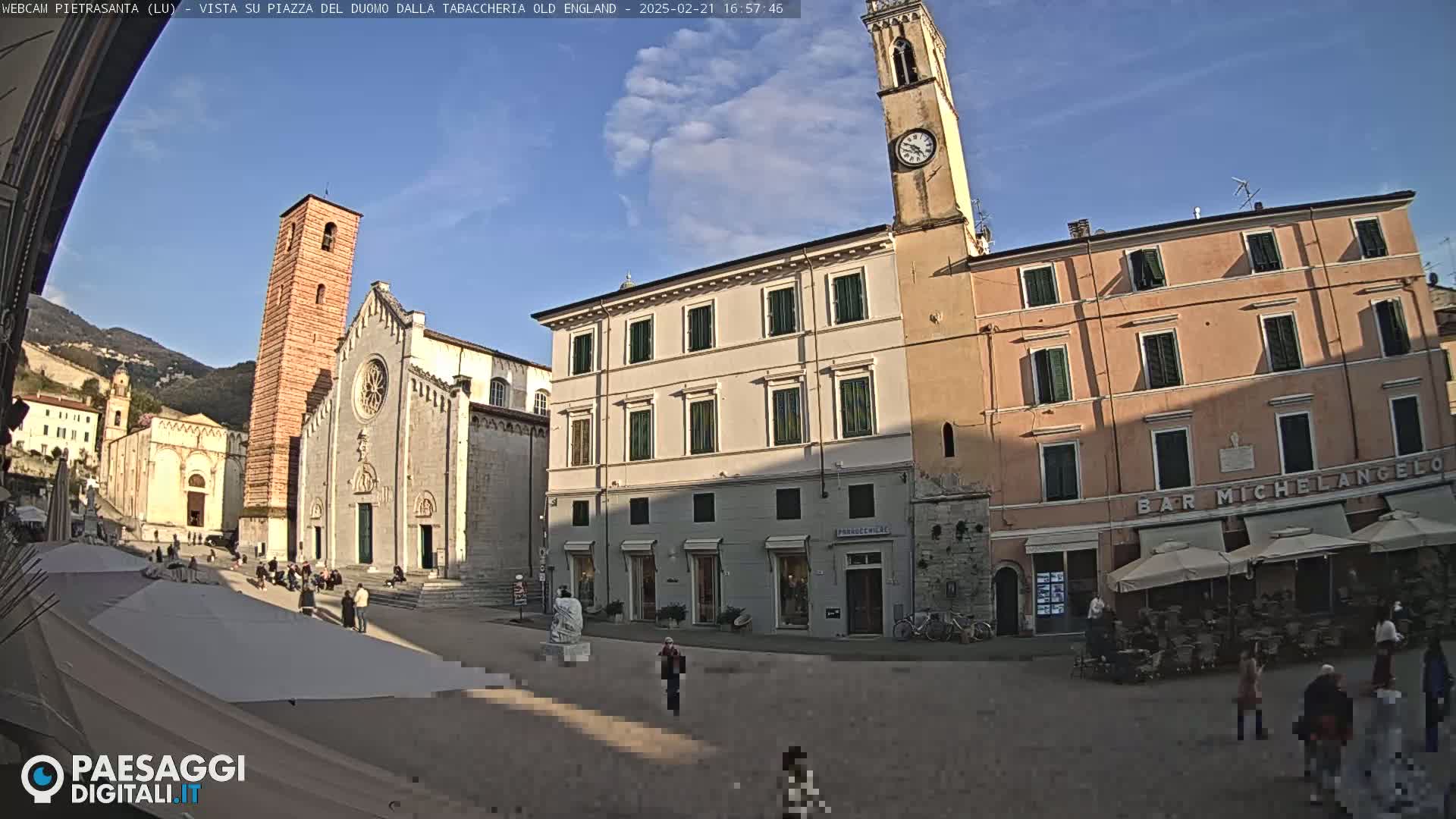 A sunny town square with a cathedral, bell tower, and several buildings, including a cafe with outdoor seating.