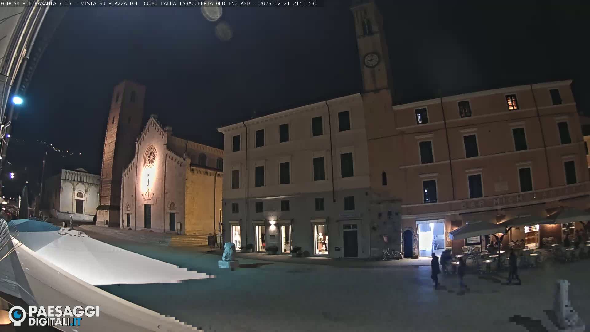 A nighttime view of a town square with a church, buildings, and a few people walking around.