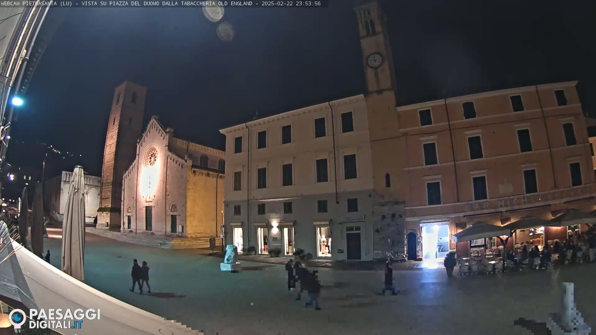 A nighttime view of a town square with a church, buildings, and people walking around under a clear sky.