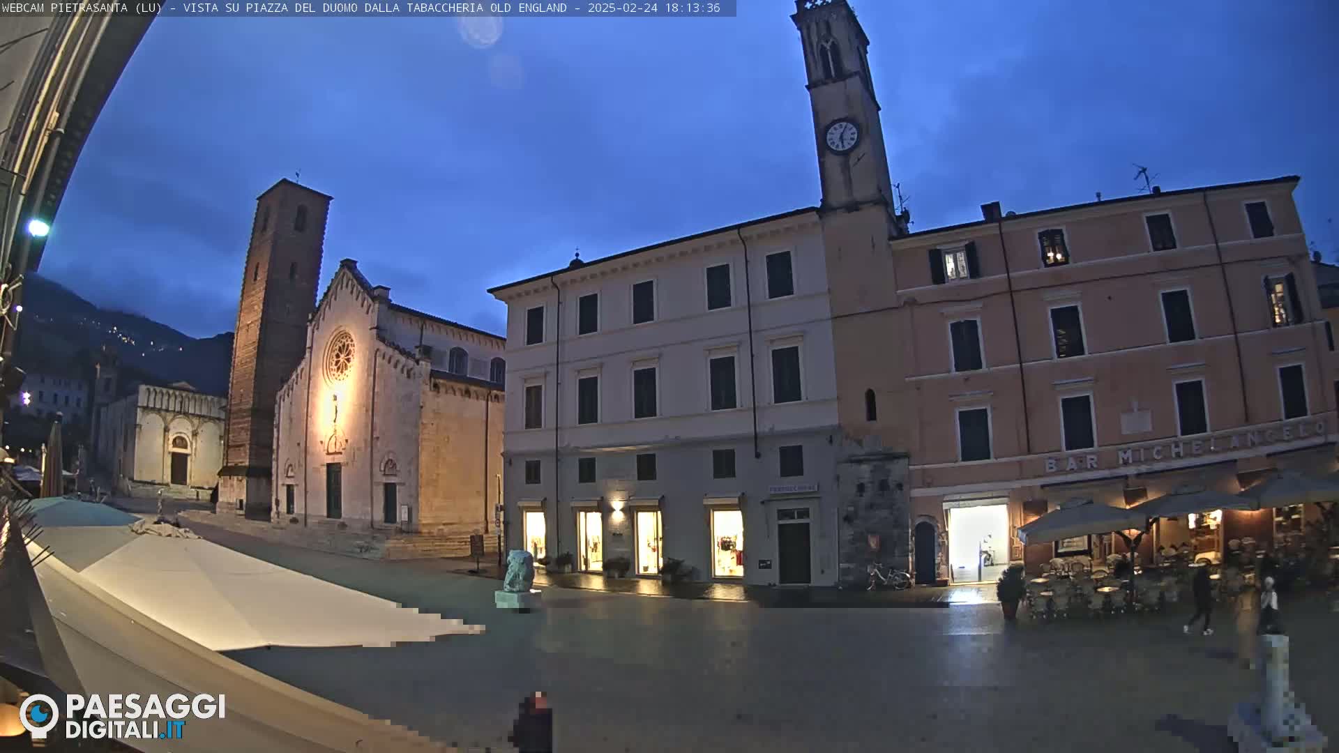 A town square at twilight, showing a church, buildings with shops, and an outdoor café under a mostly clear sky.