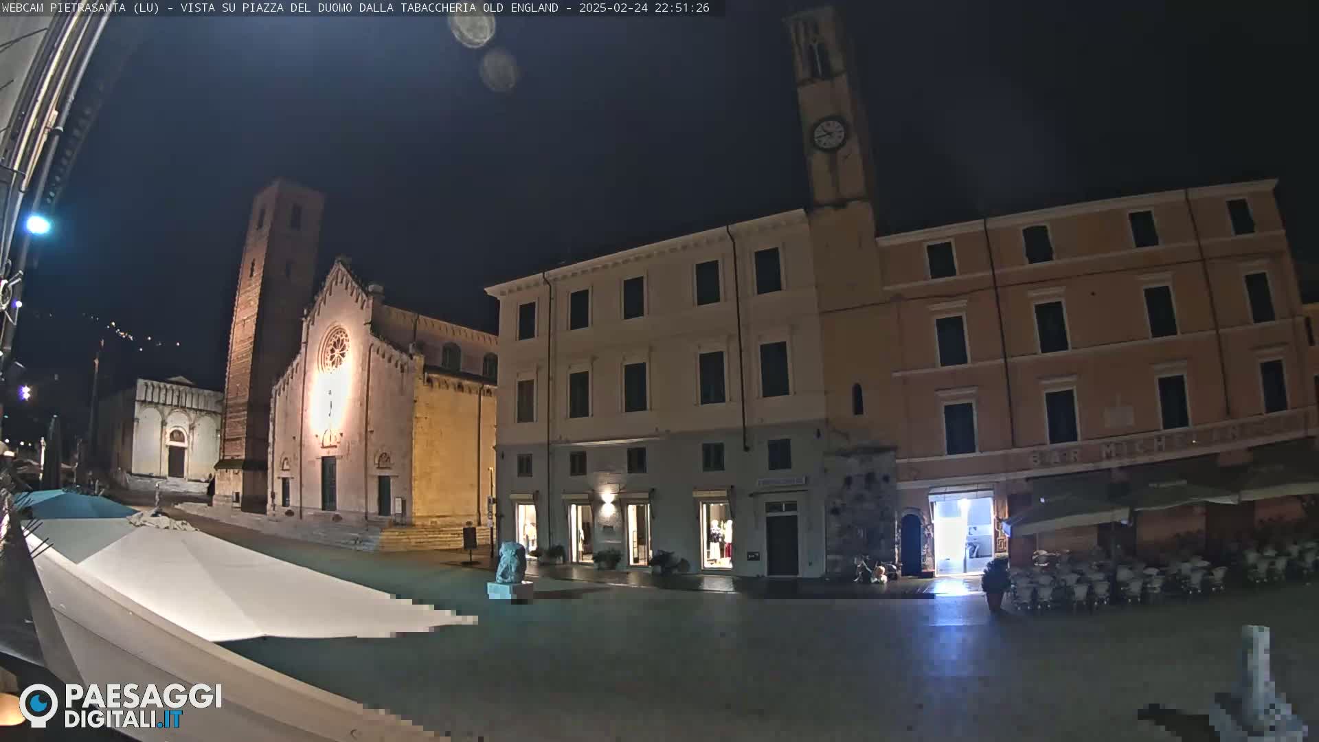 A nighttime view of a town square with a church, buildings, and a few people, under a clear, dark sky.
