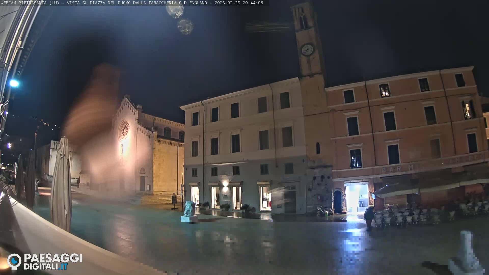 A nighttime view of a town square with illuminated buildings, a church, and a clock tower under a dark, clear sky.