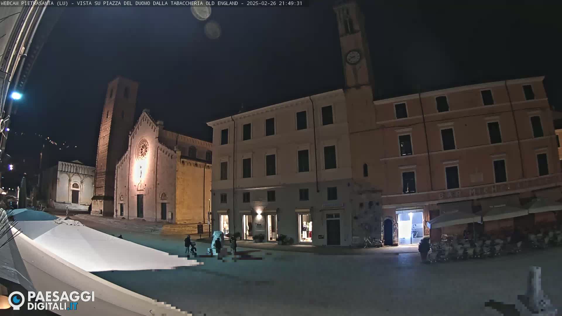 A nighttime view of a town square with a church, buildings, and a few people, under a clear, dark sky.