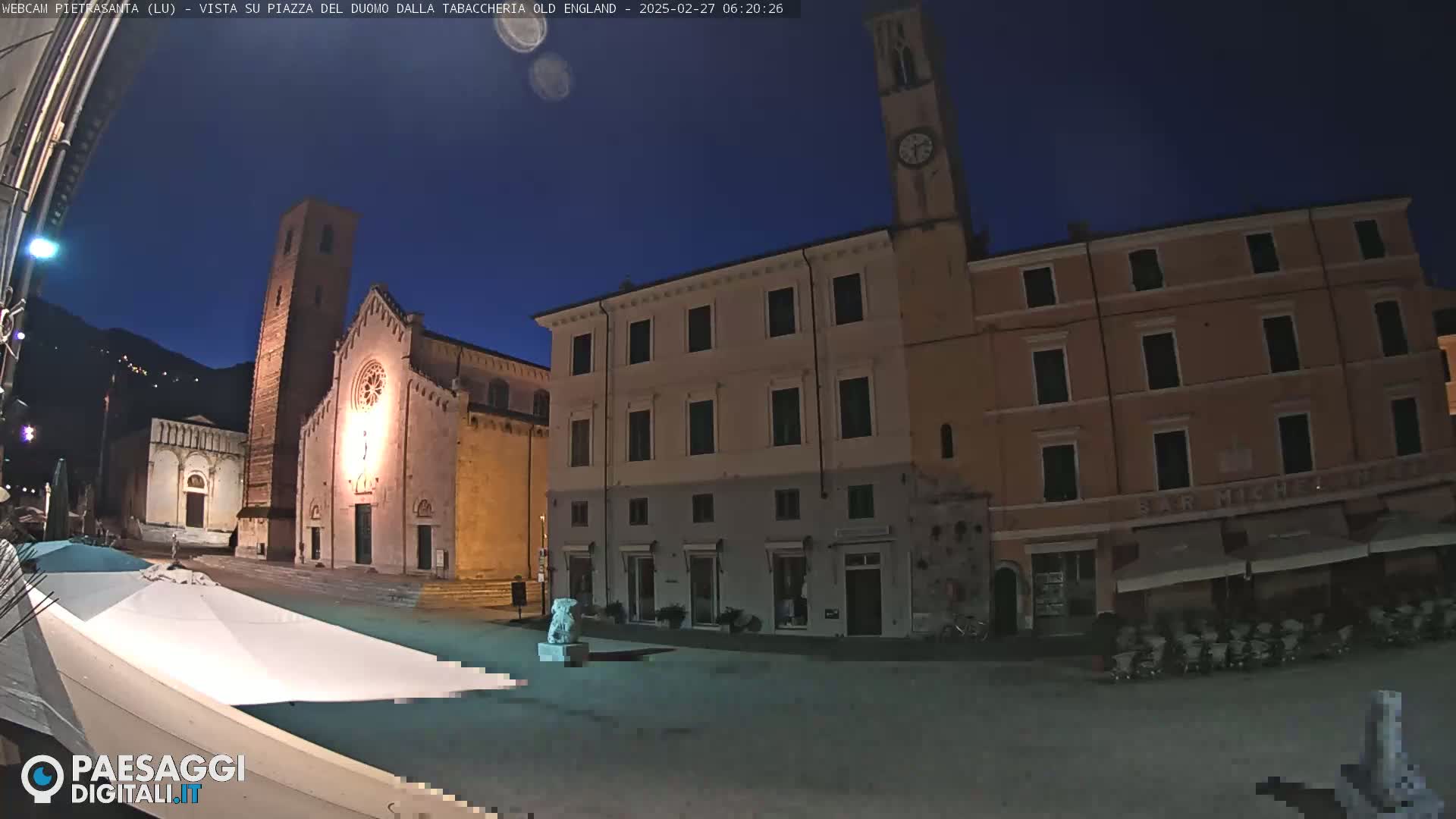 A town square at night, illuminated by artificial light, features a church, several buildings, and a statue, under a clear, dark blue twilight sky.