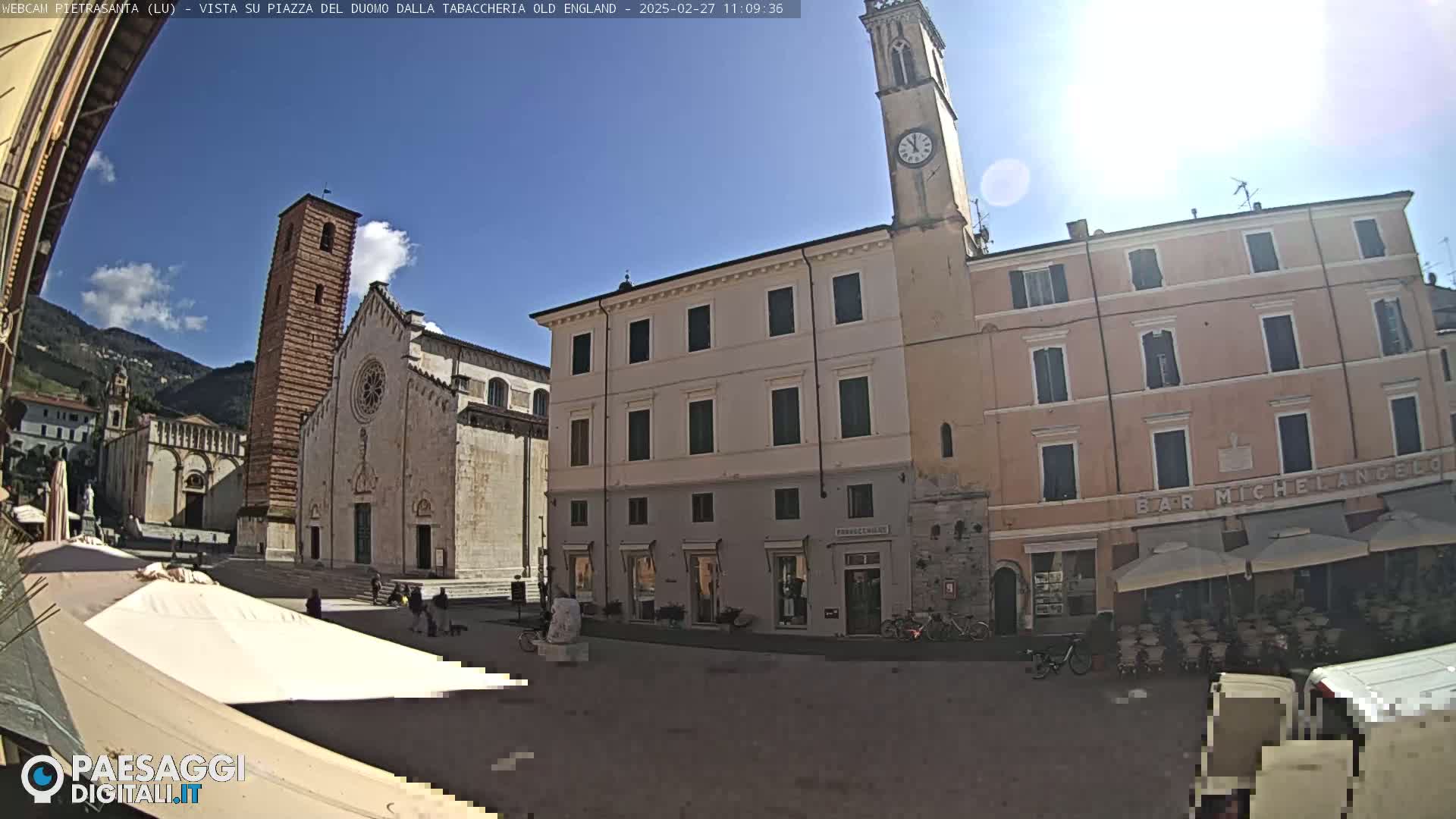 A sunny day reveals a town square with a large cathedral, several buildings, and outdoor seating areas.
