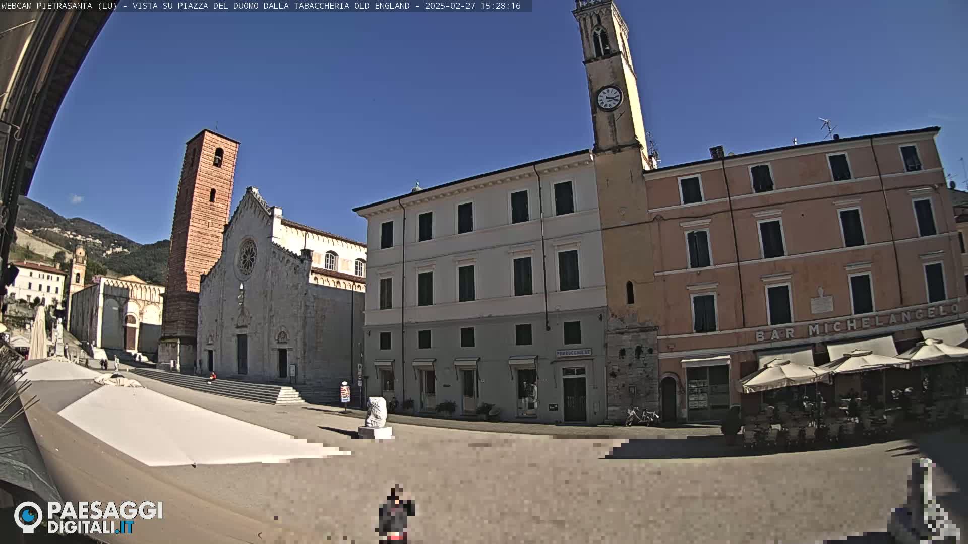 A sunny town square is shown, featuring a large cathedral with a tall bell tower, adjacent buildings, and outdoor café seating.