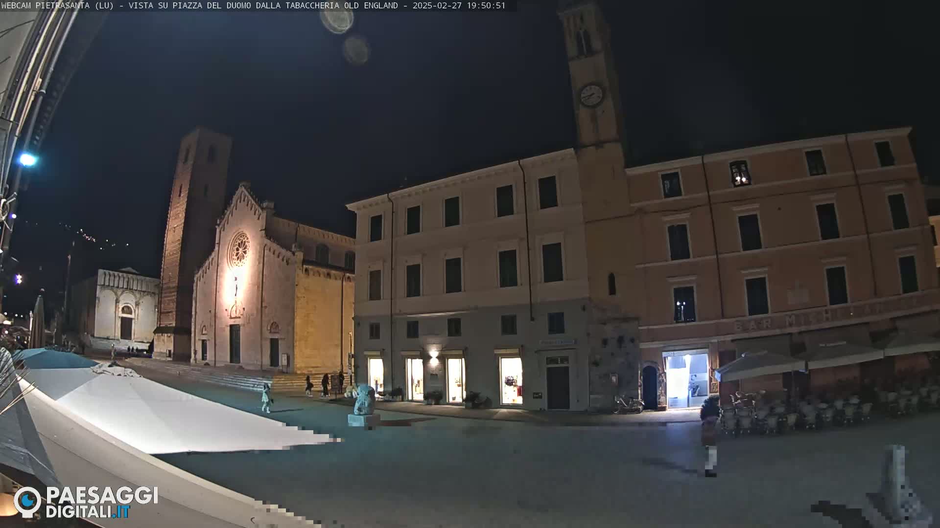 A nighttime view of a town square with a church, buildings, and a few people, under a dark, clear sky.