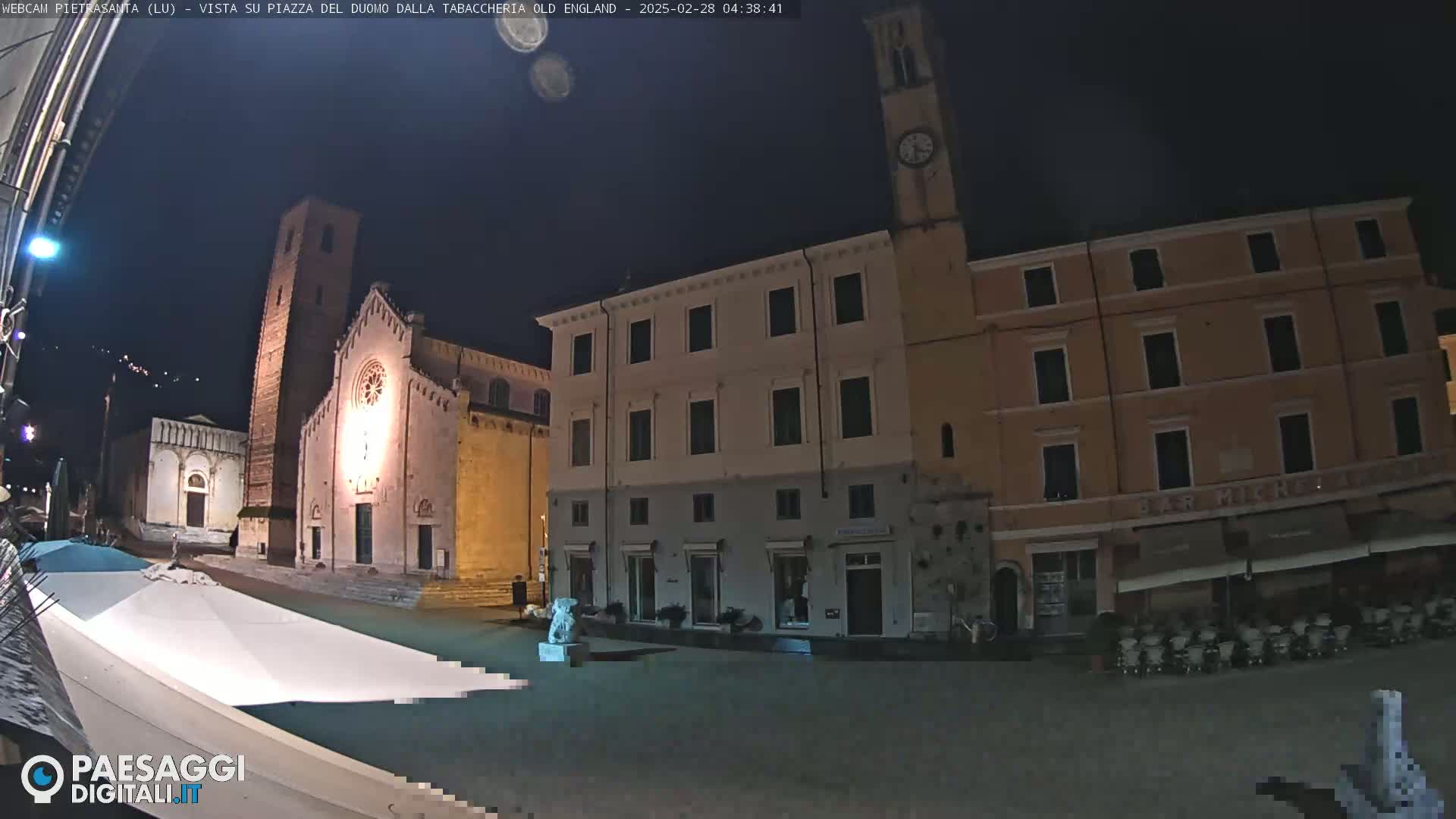 A nighttime view of a town square with a church, buildings, and a clock tower under a clear, dark sky.