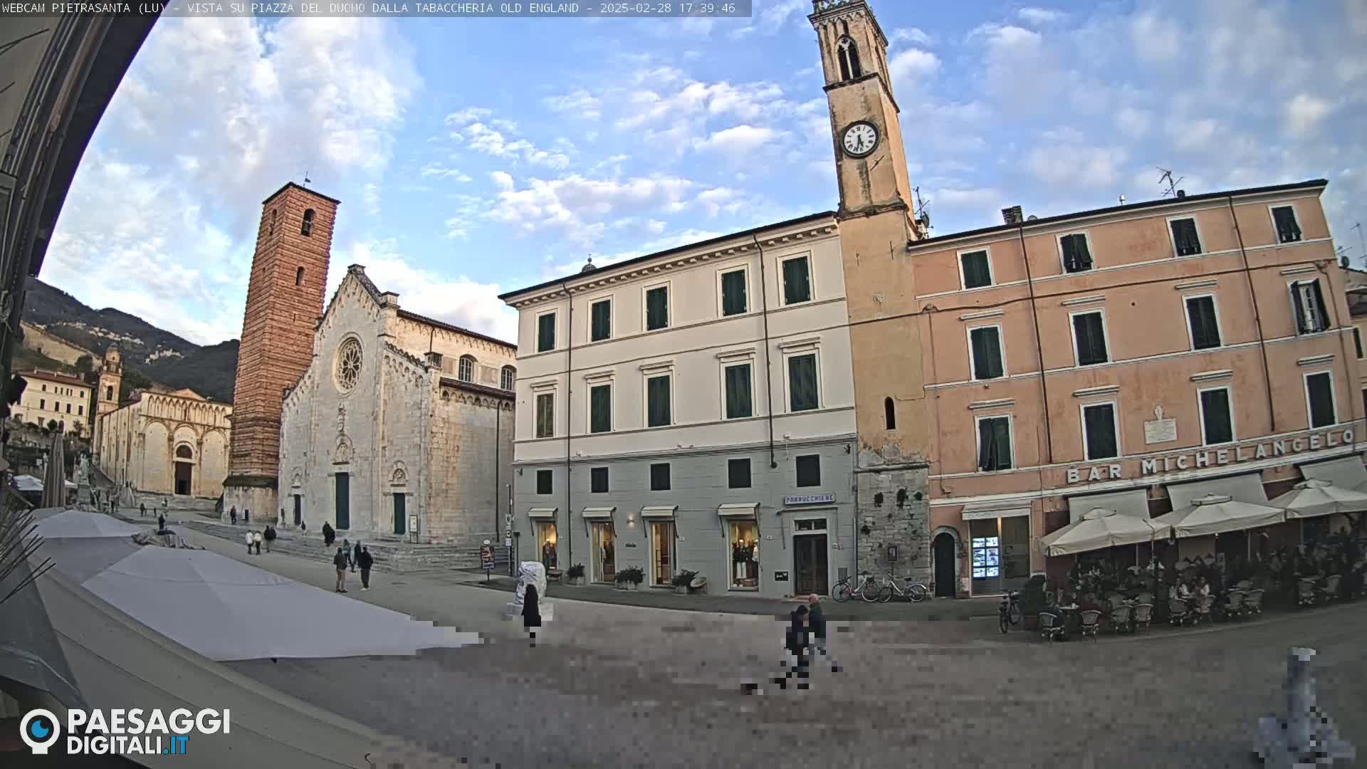 A town square with a large church, several buildings, and an outdoor cafe under a partly cloudy sky.